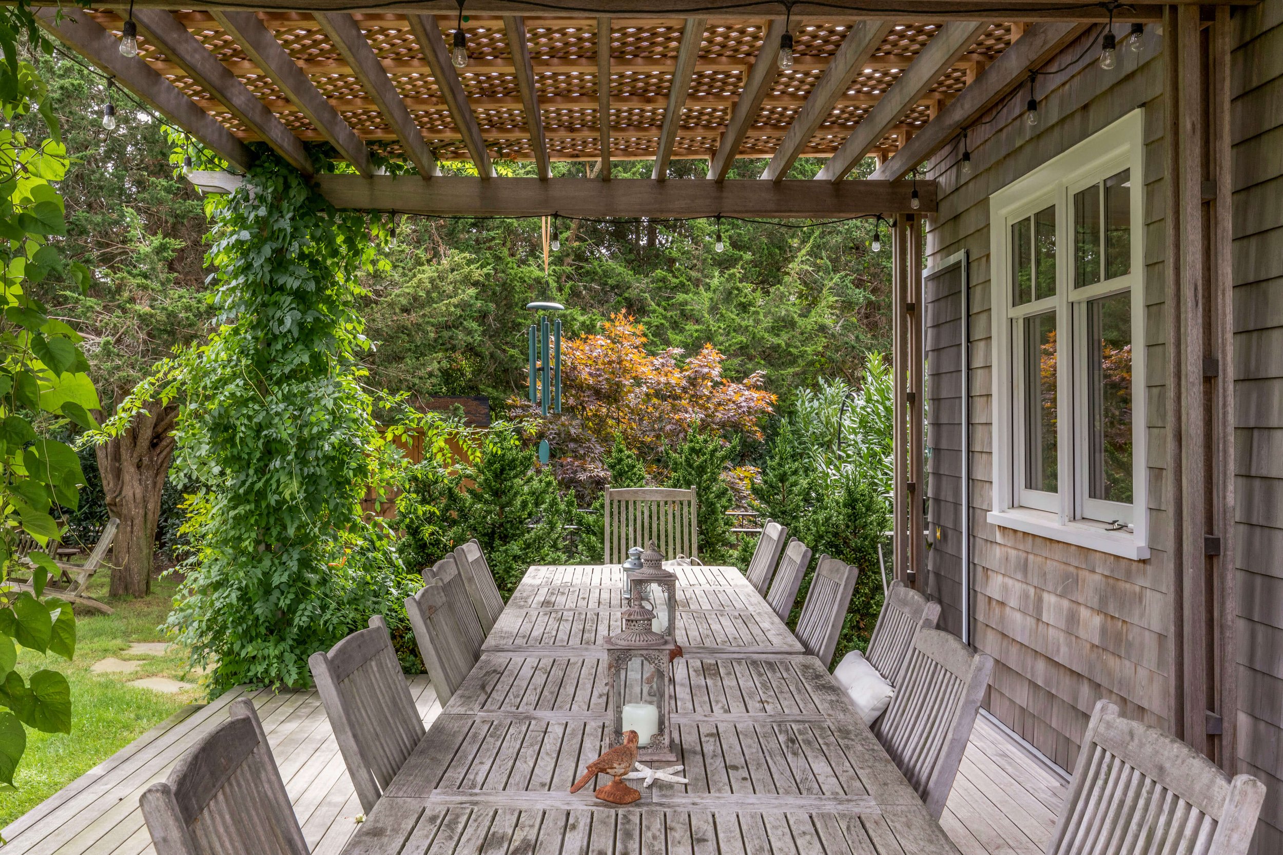 Outdoor wooden dining table under a pergola, surrounded by greenery and lanterns on the table.