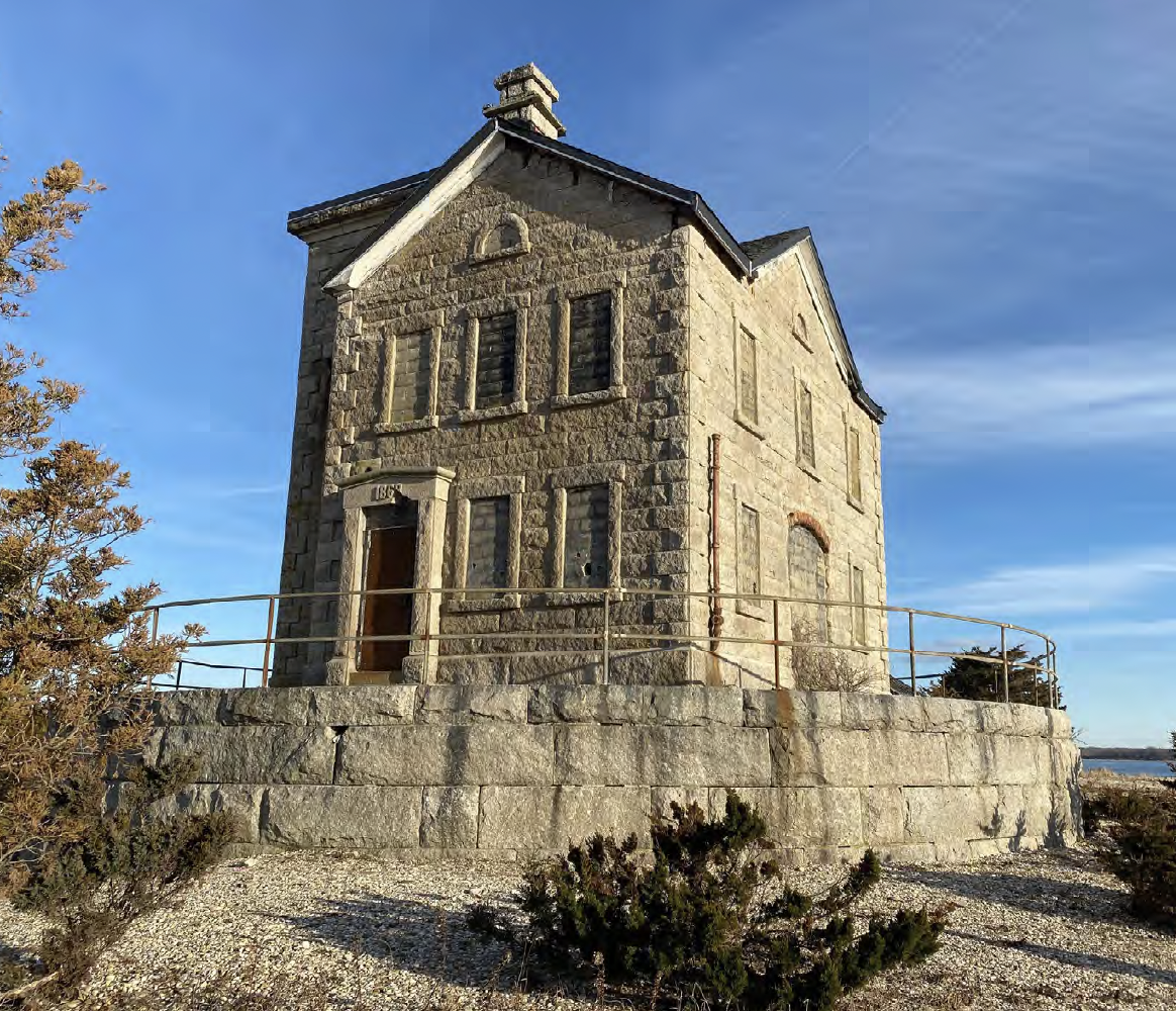 Historic stone building with arched windows and door, surrounded by a fence, against a clear blue sky.