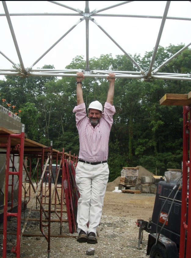 Person in a hard hat and striped shirt hanging from metal scaffolding at a construction site with trees in the background.