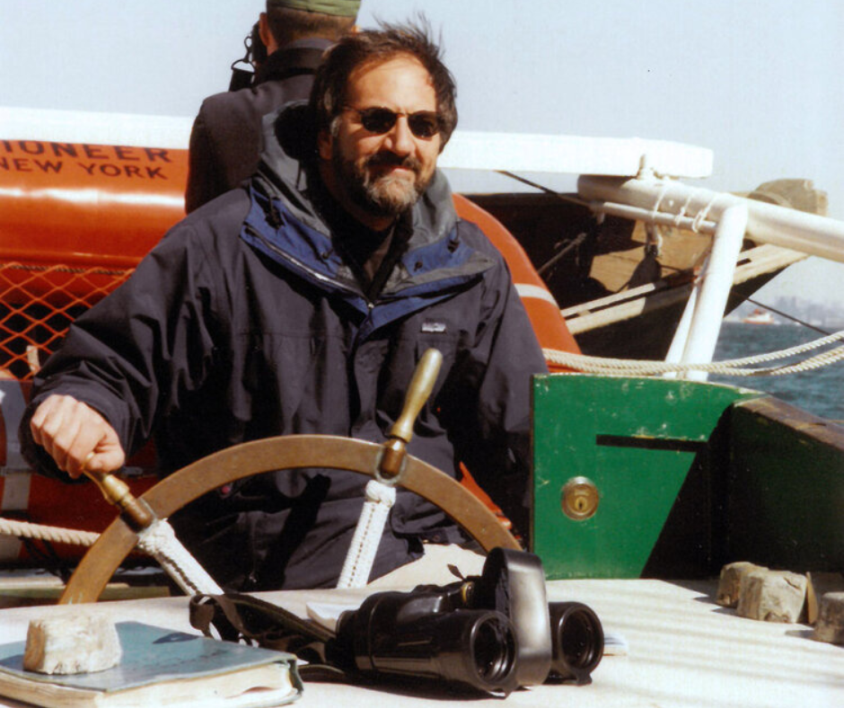 Person steering a boat using a large wooden wheel, with binoculars and a book on the deck.
