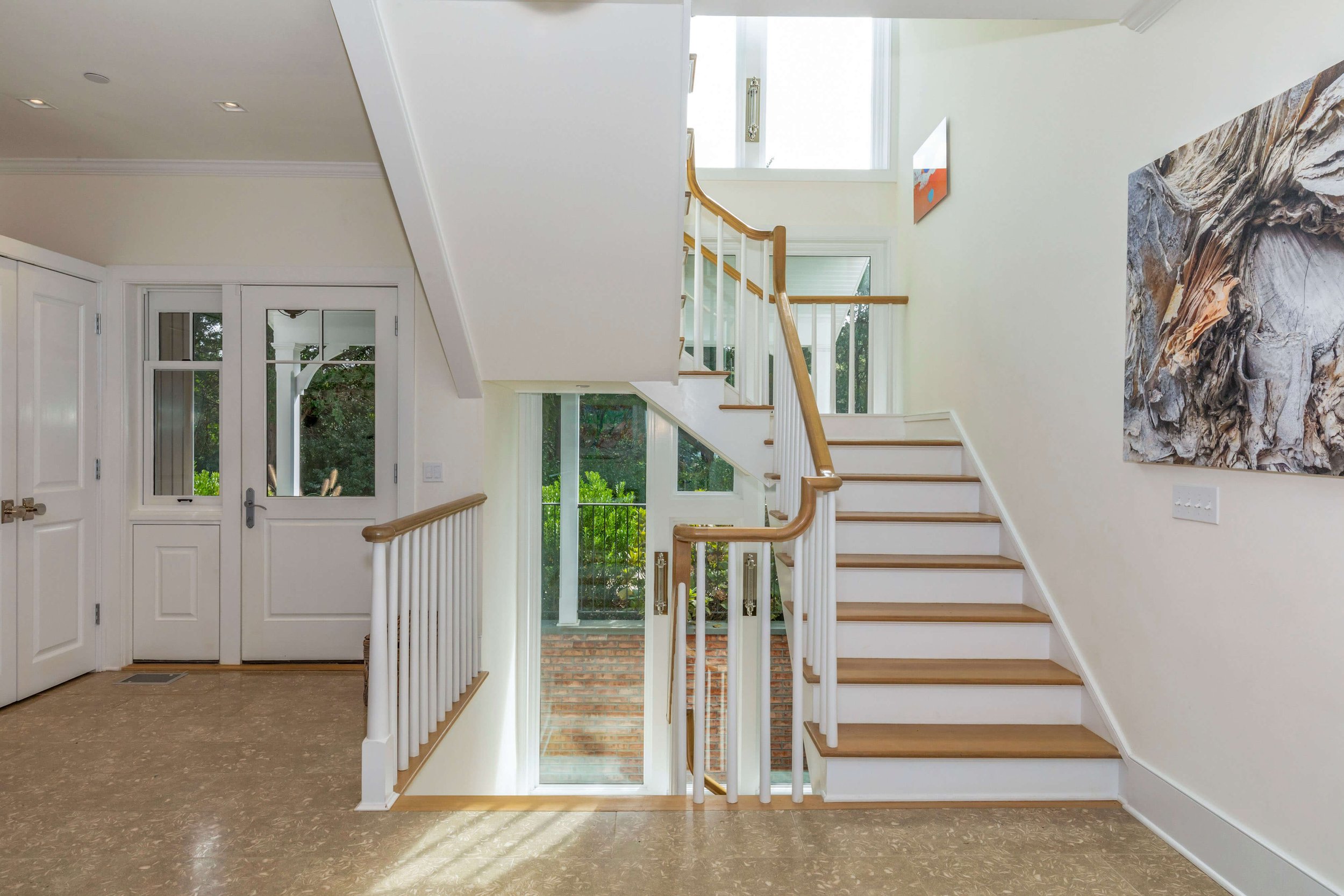 Bright entryway with staircase, glass front door, and large windows, featuring two artworks on cream-colored walls.