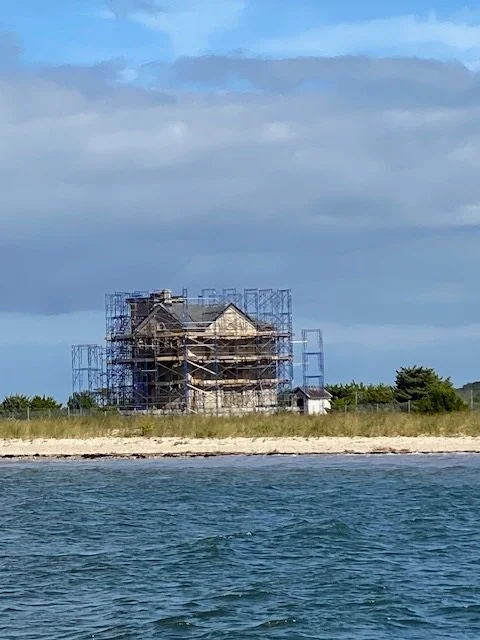 House under construction surrounded by scaffolding near a shoreline, with grass and water in the foreground.
