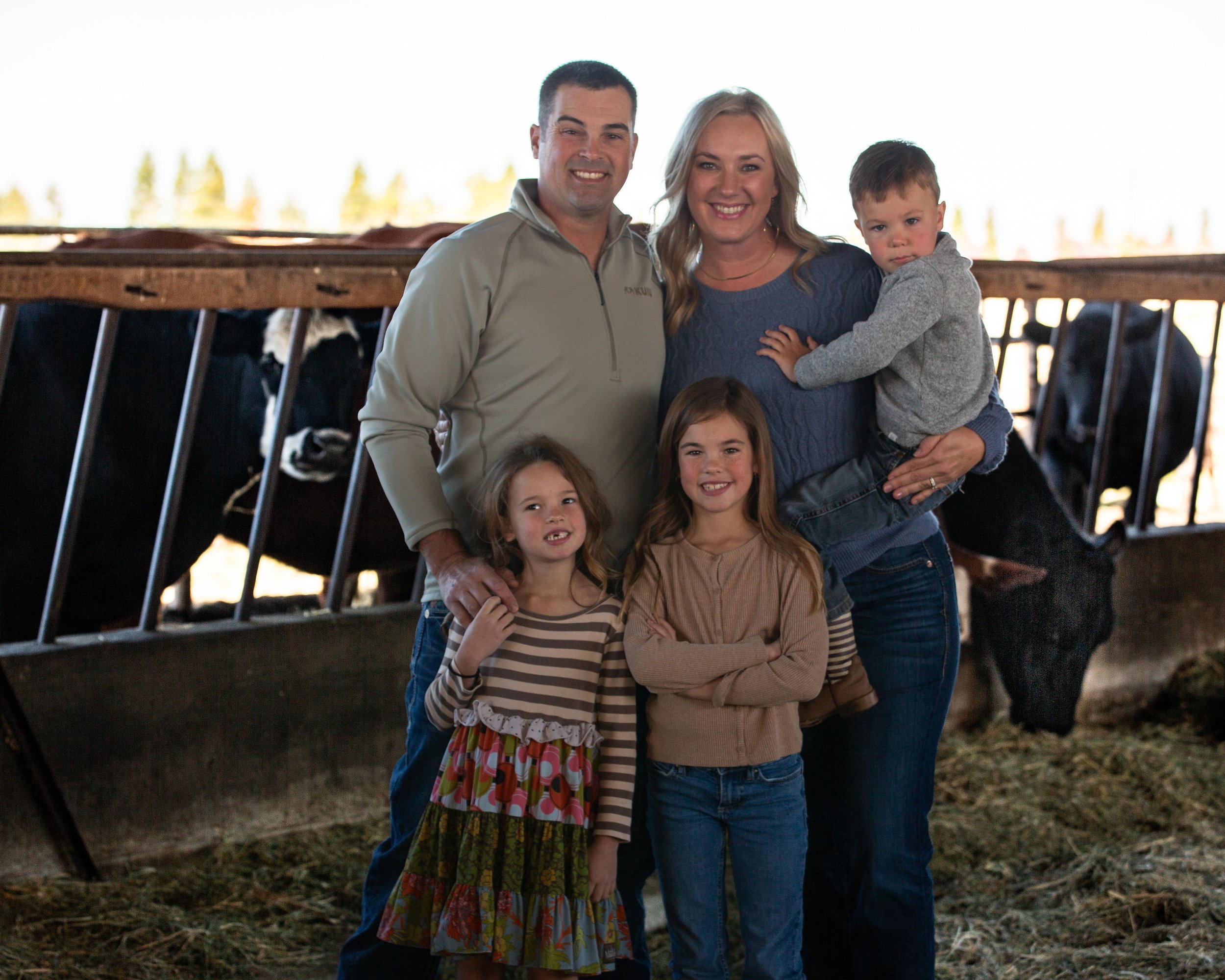 A family of five standing together inside a barn with cows in the background. The father and mother are smiling, with the father wearing a light gray zip-up shirt and the mother wearing a blue sweater. Three children are with them: a girl in a colorf