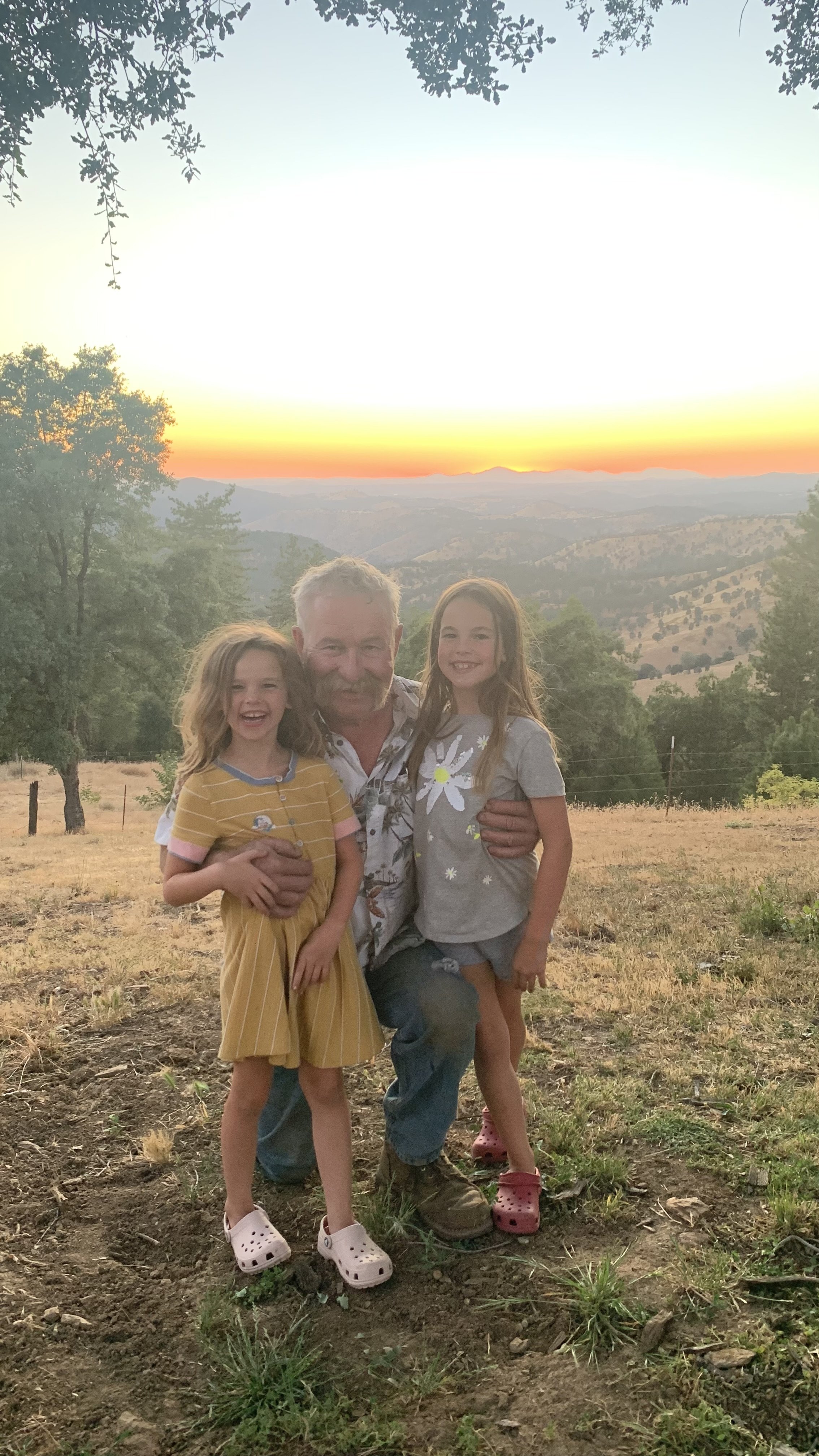 A man with two young girls smiling and hugging during sunset in a rural outdoor setting with trees and distant hills.