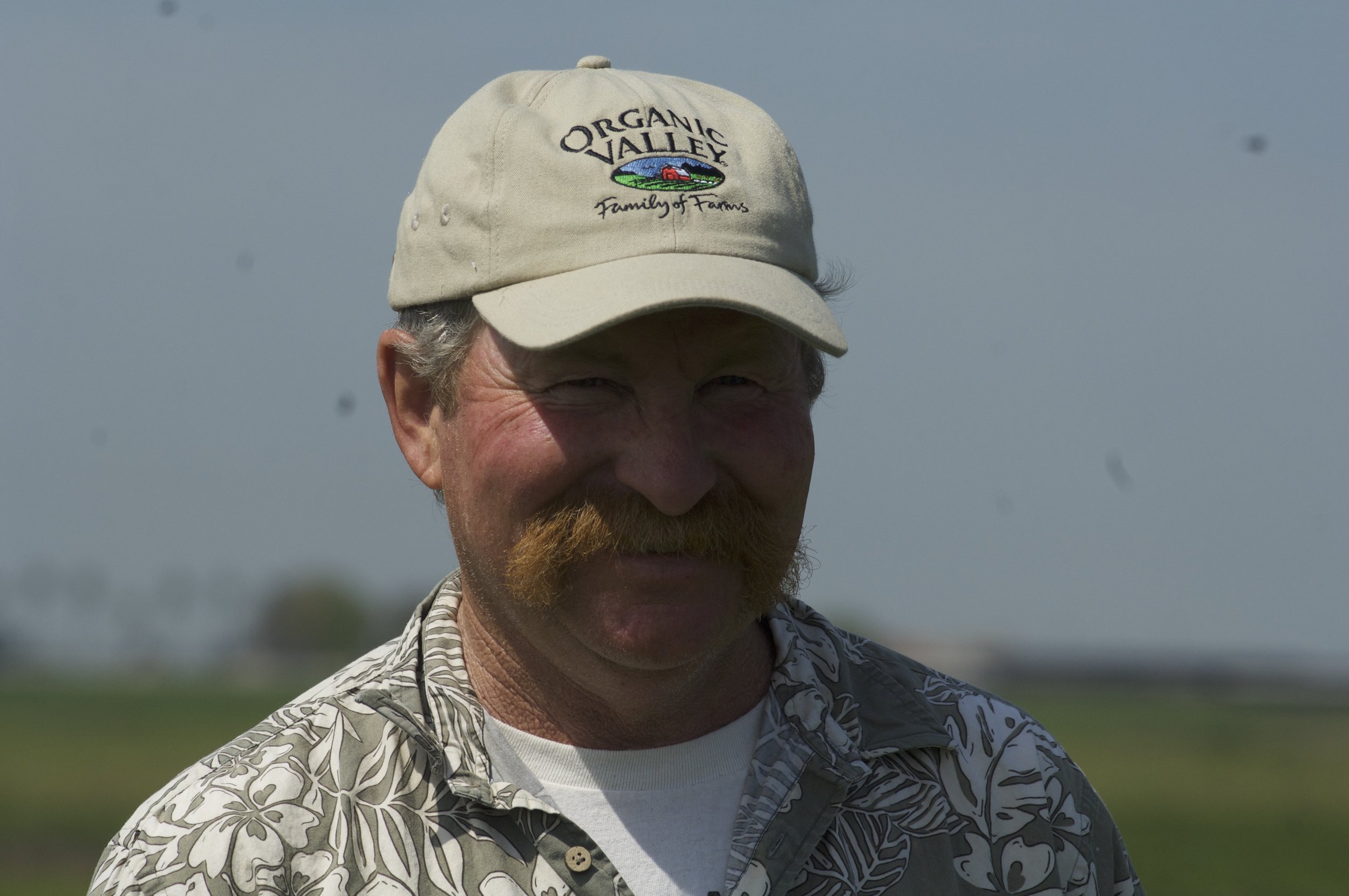 A man with a red beard wearing a beige cap with the words 'Organic Valley' and a logo, and a patterned shirt, outdoors on a sunny day.