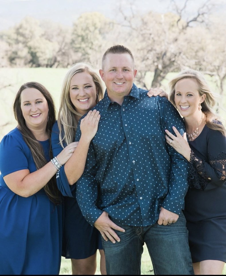 Four people, three women and one man, smiling outdoors in a park or field with trees in the background. They are dressed in shades of blue and appear to be posing for a photo.