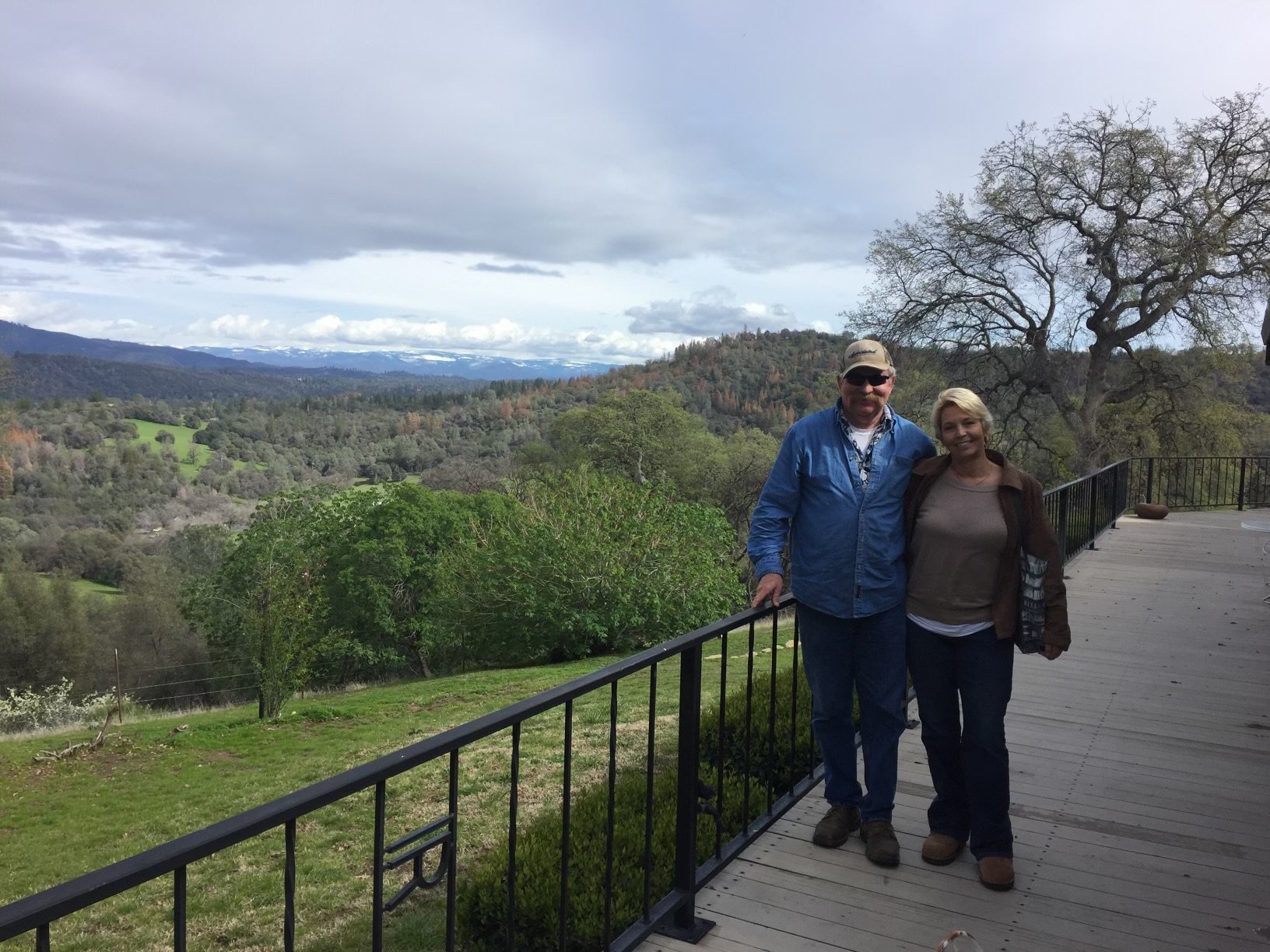 Two people standing on a wooden balcony with a scenic view of green hills, trees, and distant mountains under a cloudy sky.