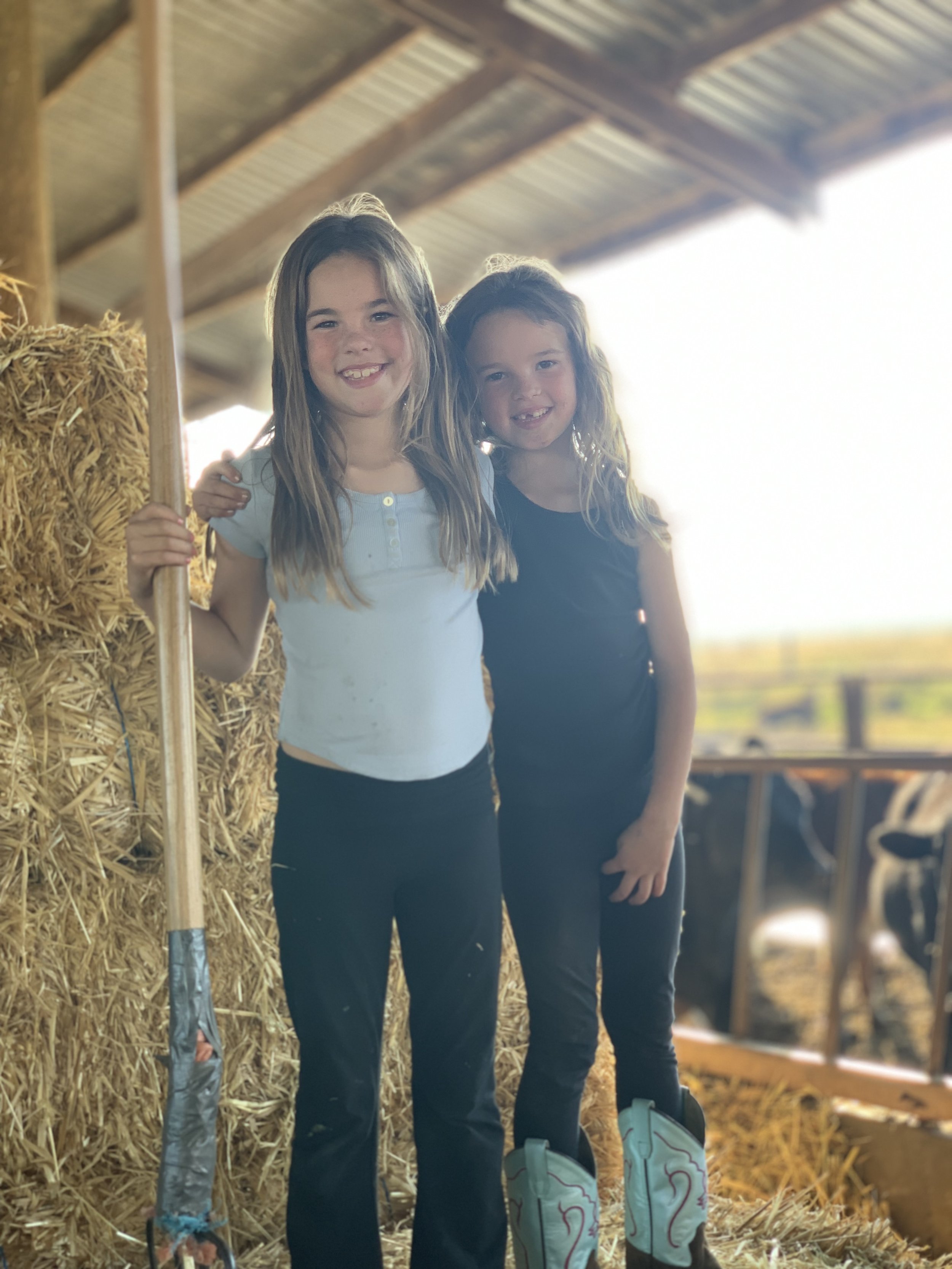 Two young girls standing in a barn with hay bales, smiling at the camera.