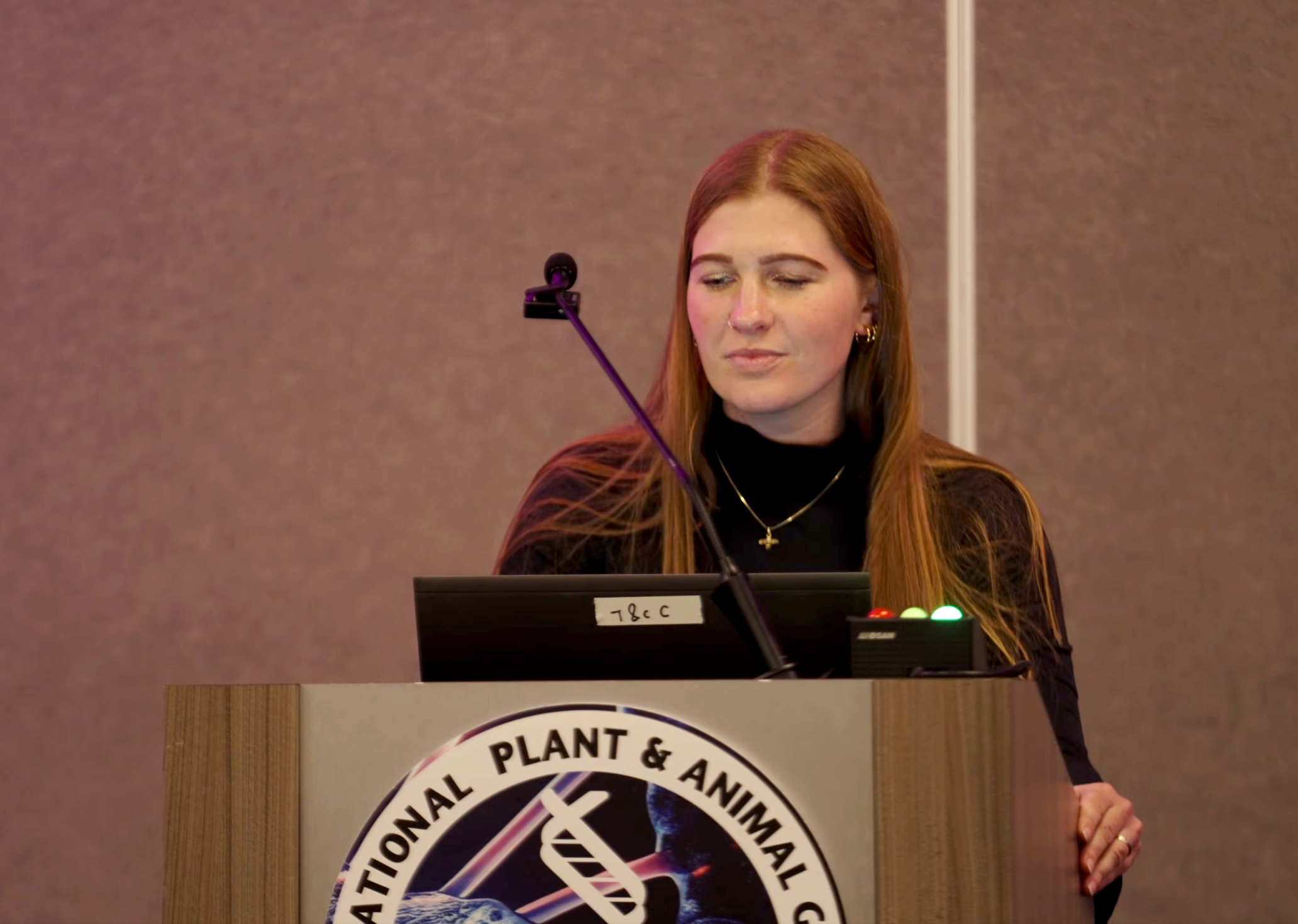 A woman with long red hair standing behind a decorated podium, appearing to give a speech, in a conference room with brown walls.