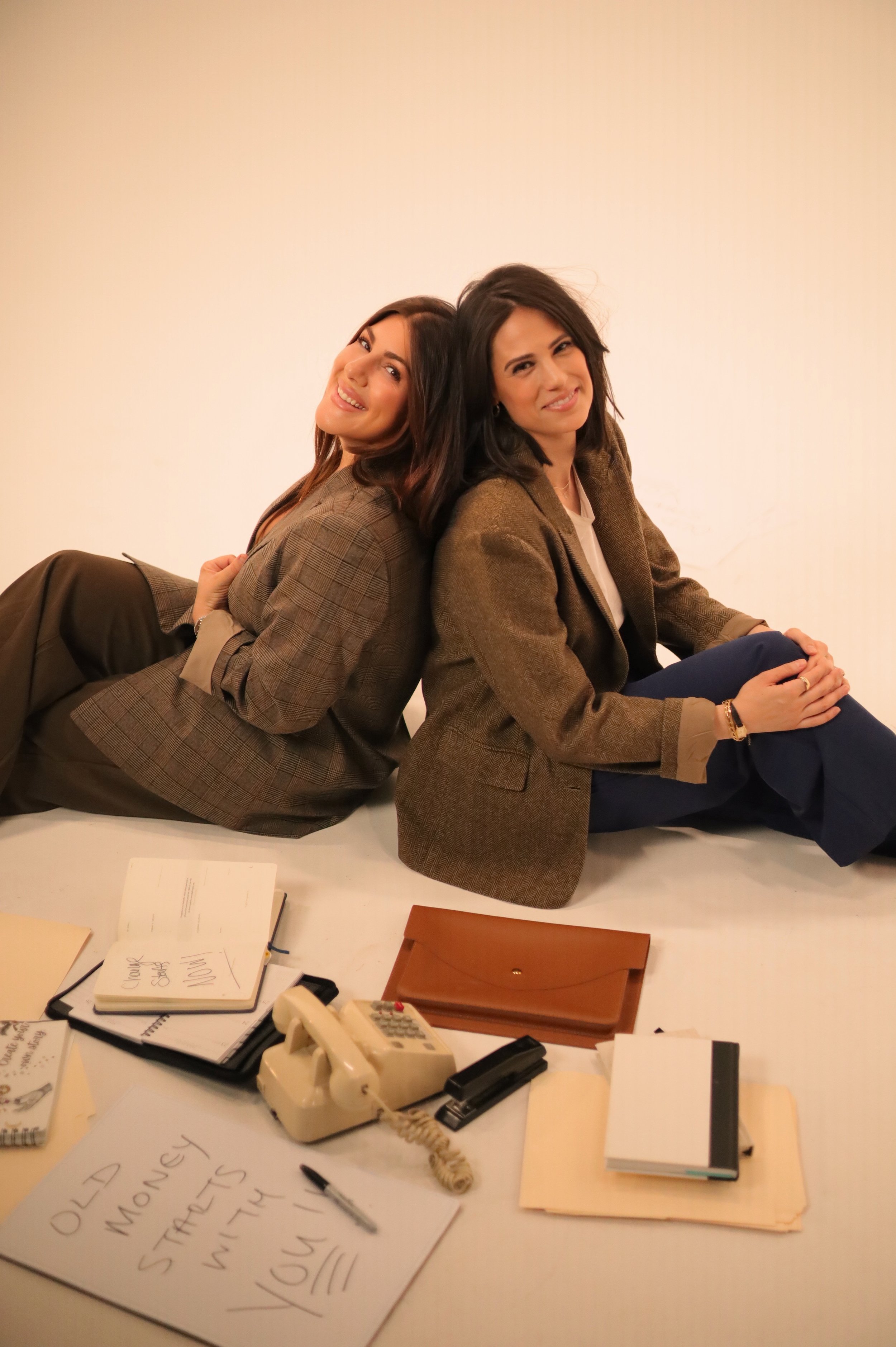 Two women sit back-to-back on a table, smiling and looking at the camera. They wear business blazers. In front of them are office supplies, notebooks, a calculator, and a sign that reads 'Old Money Strategies with You!'