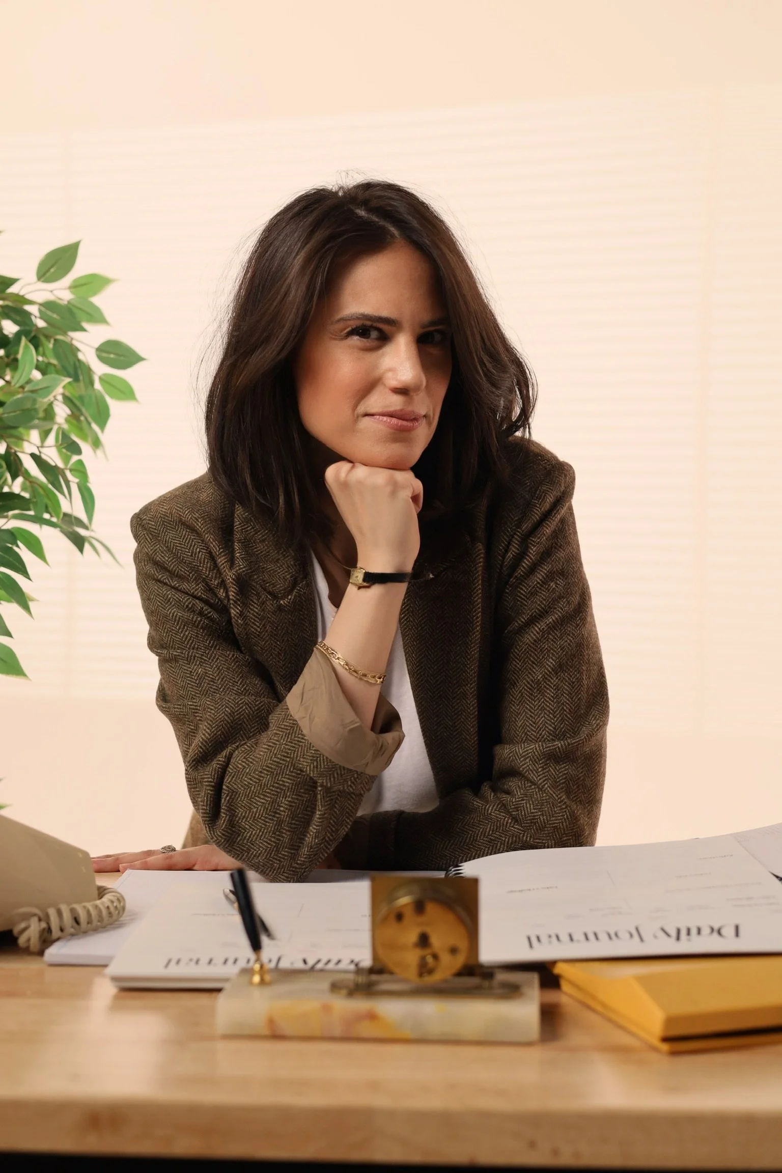 A woman with shoulder-length brown hair sitting at a desk, resting her chin on her hand, wearing a brown blazer and gold jewelry, with a desk clock and papers in front of her, and a plant to her left.