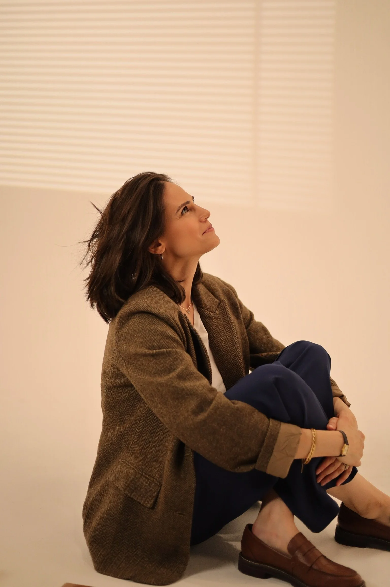 A woman sitting on the floor, looking up thoughtfully with a neutral background.
