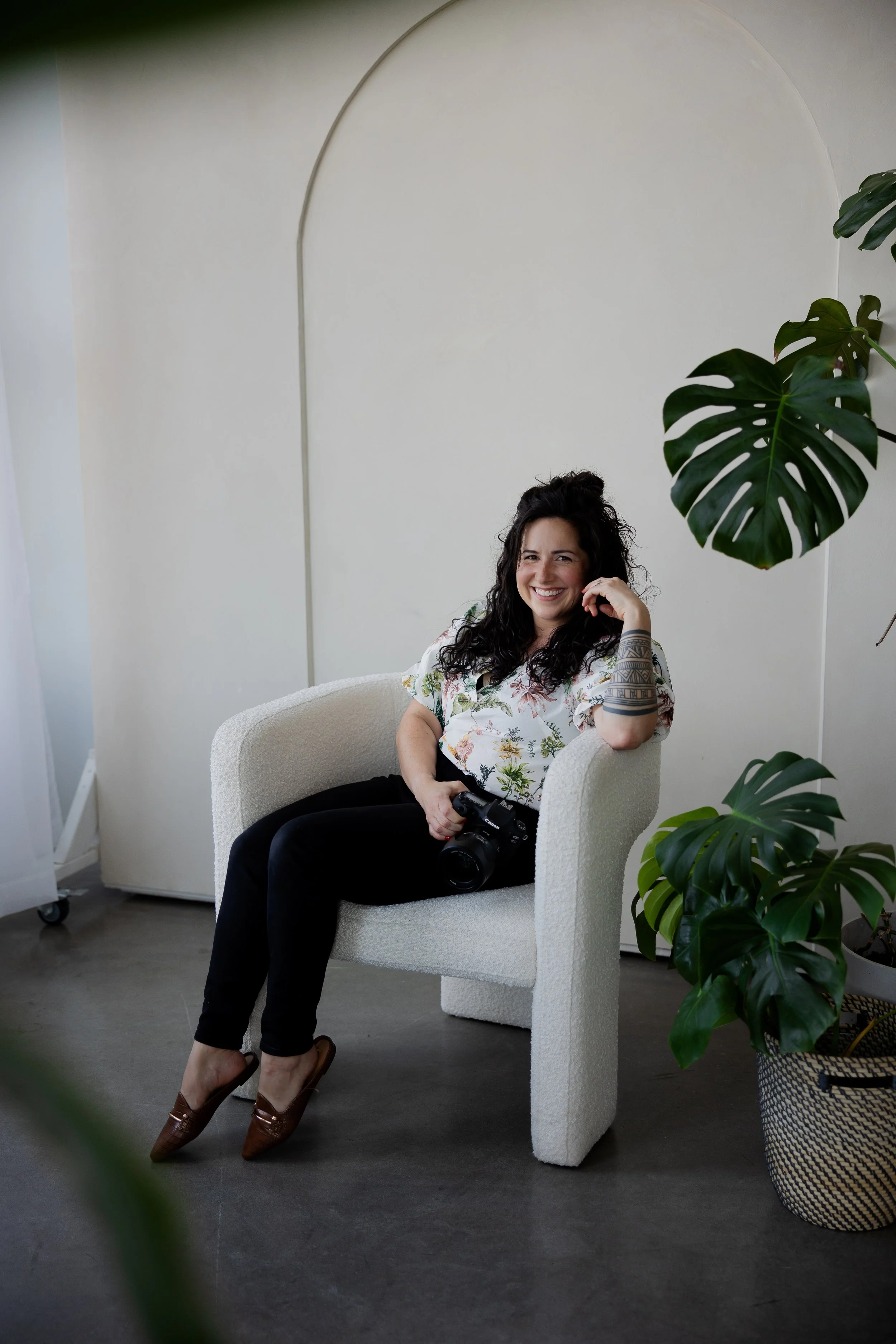 A woman with dark, curly hair smiling while sitting in a beige armchair, holding a camera in her lap, in a room with white walls and large green houseplants.