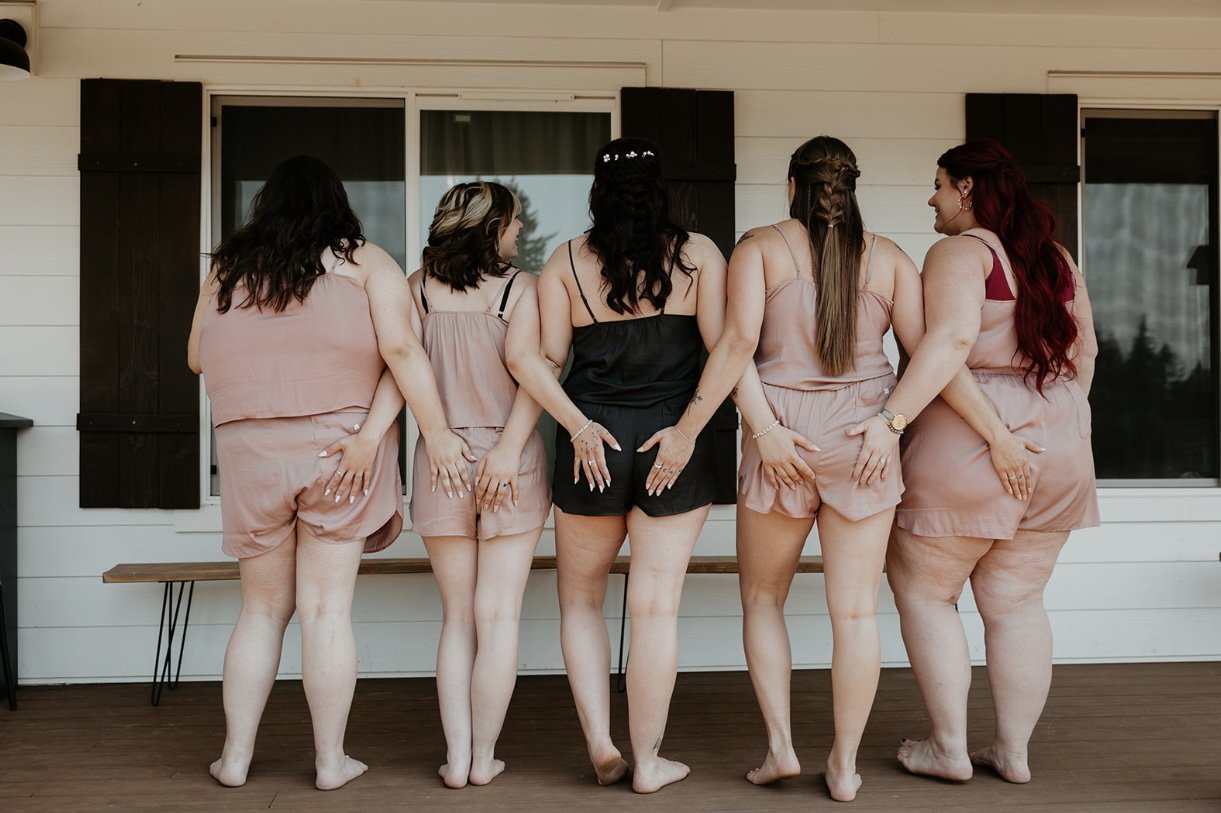 Five women standing barefoot on a wooden porch, showing the back view with their hands on their hips or behind their backs, wearing matching pink and black lounge outfits, in front of a white house with black shutters.
