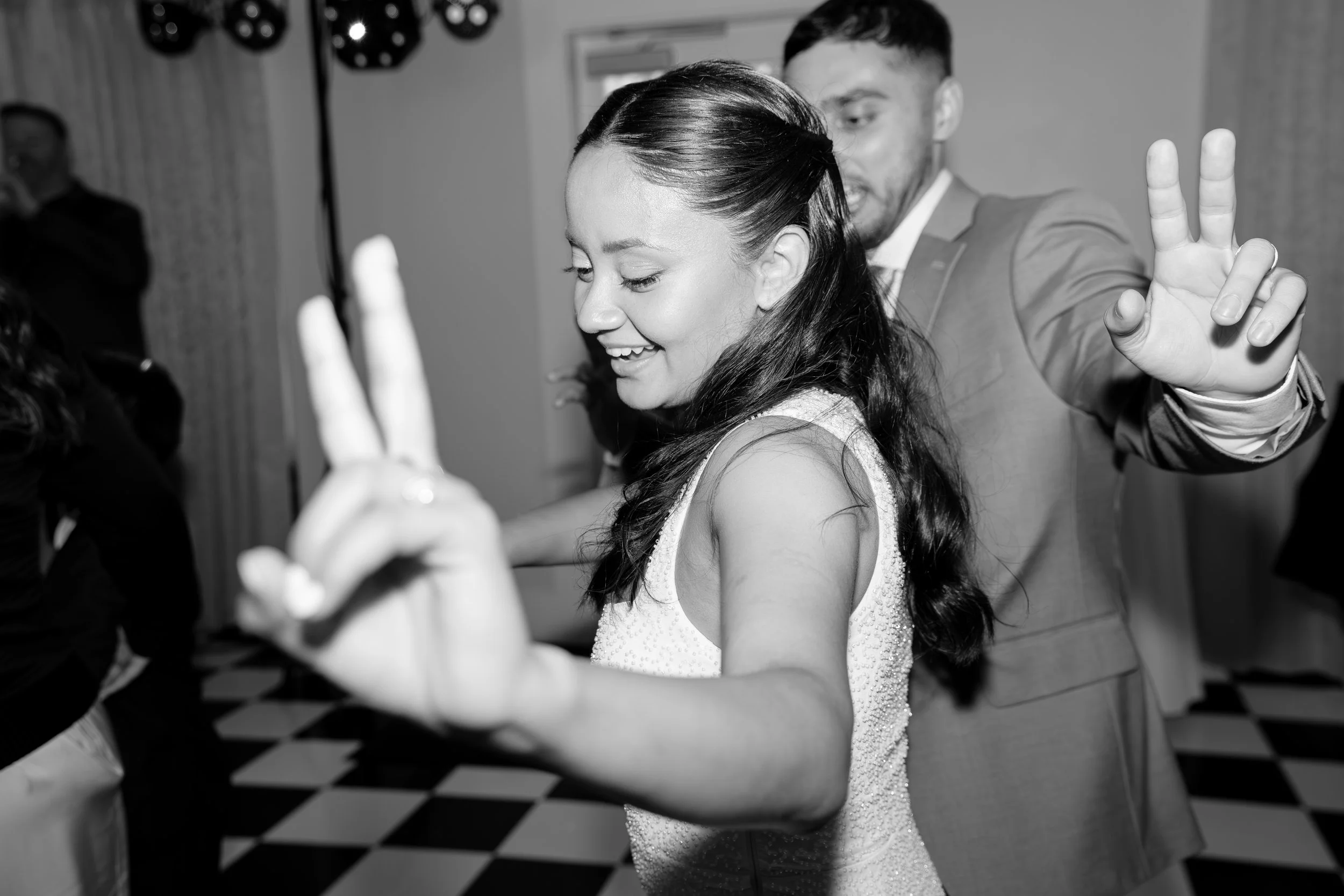 A woman with long dark hair and a sparkly dress is dancing with a man in a suit at a party, both smiling and making peace signs with their hands. Wedding reception dance floor photos. Flash photography. Traditional Indian wedding photographer