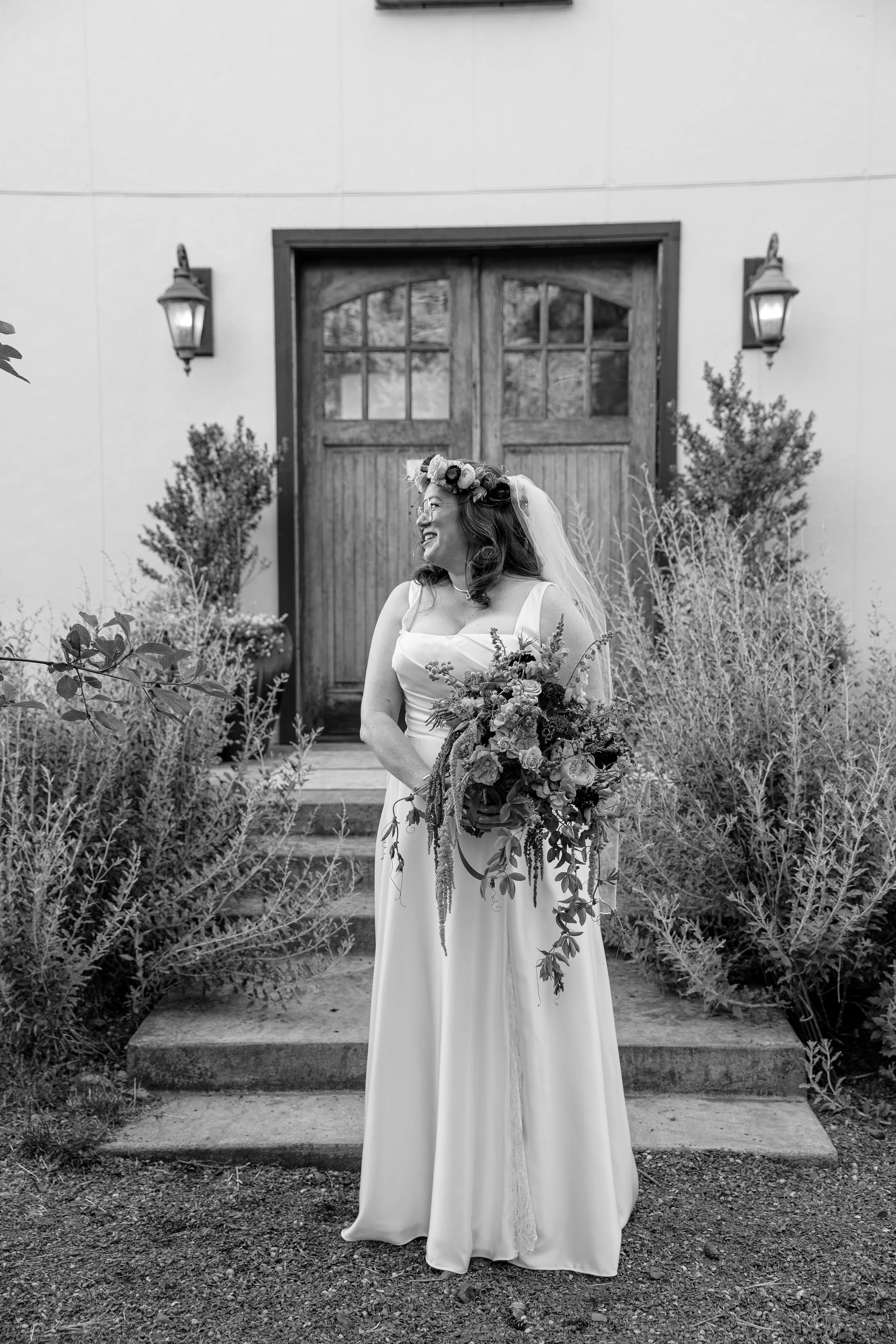 Black and white photo of a bride standing on steps in front of a wooden double door with window panes. She is wearing a wedding dress, a veil, glasses, and a floral crown, and holding a large bouquet of flowers. Oregon + Travel Wedding Photographer
