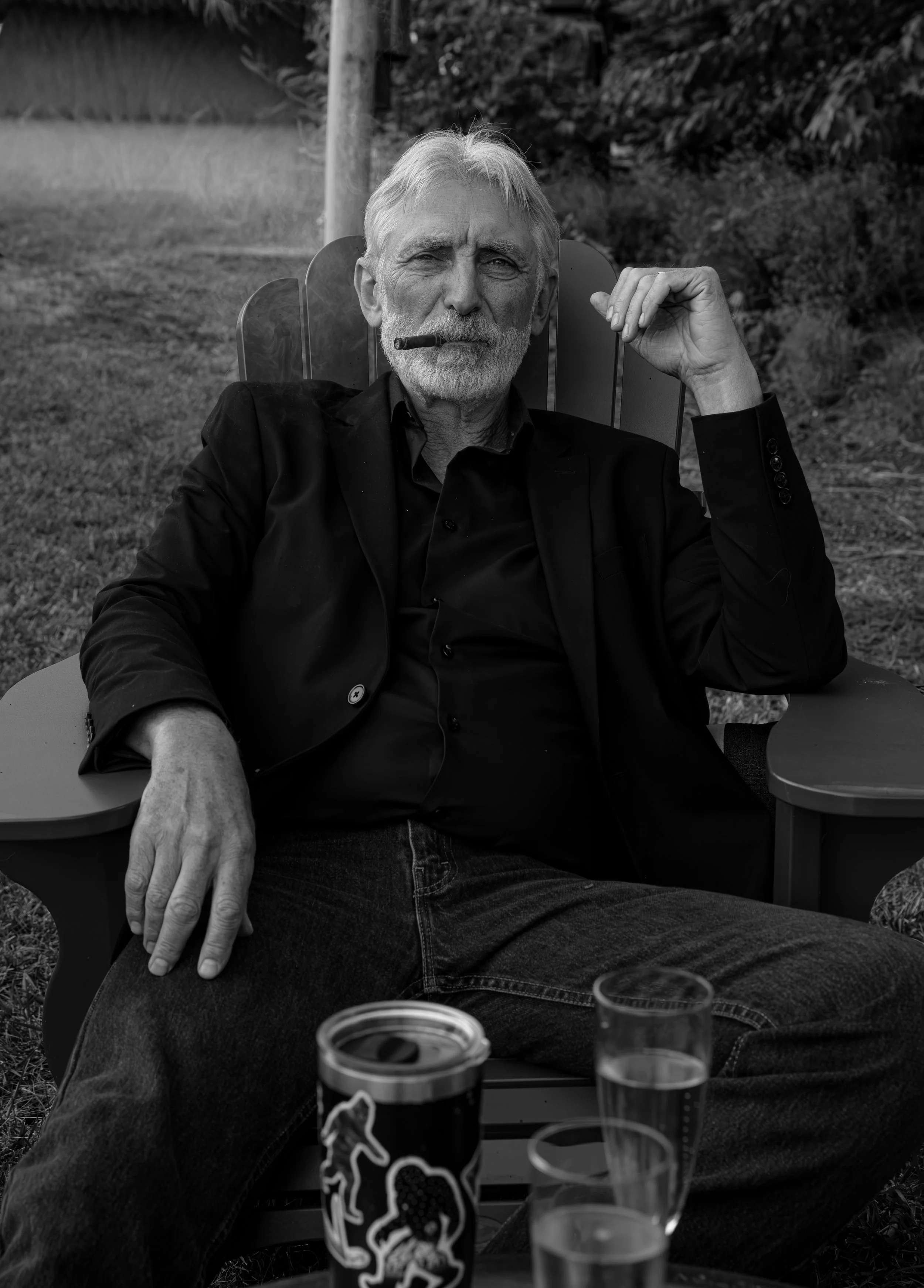 An older man with gray hair and beard is sitting outdoors on a chair, smoking a cigar, and wearing a black jacket. Portrait photography in Portland Oregon California Arizona New York Utah and worldwide. Wedding guest photos