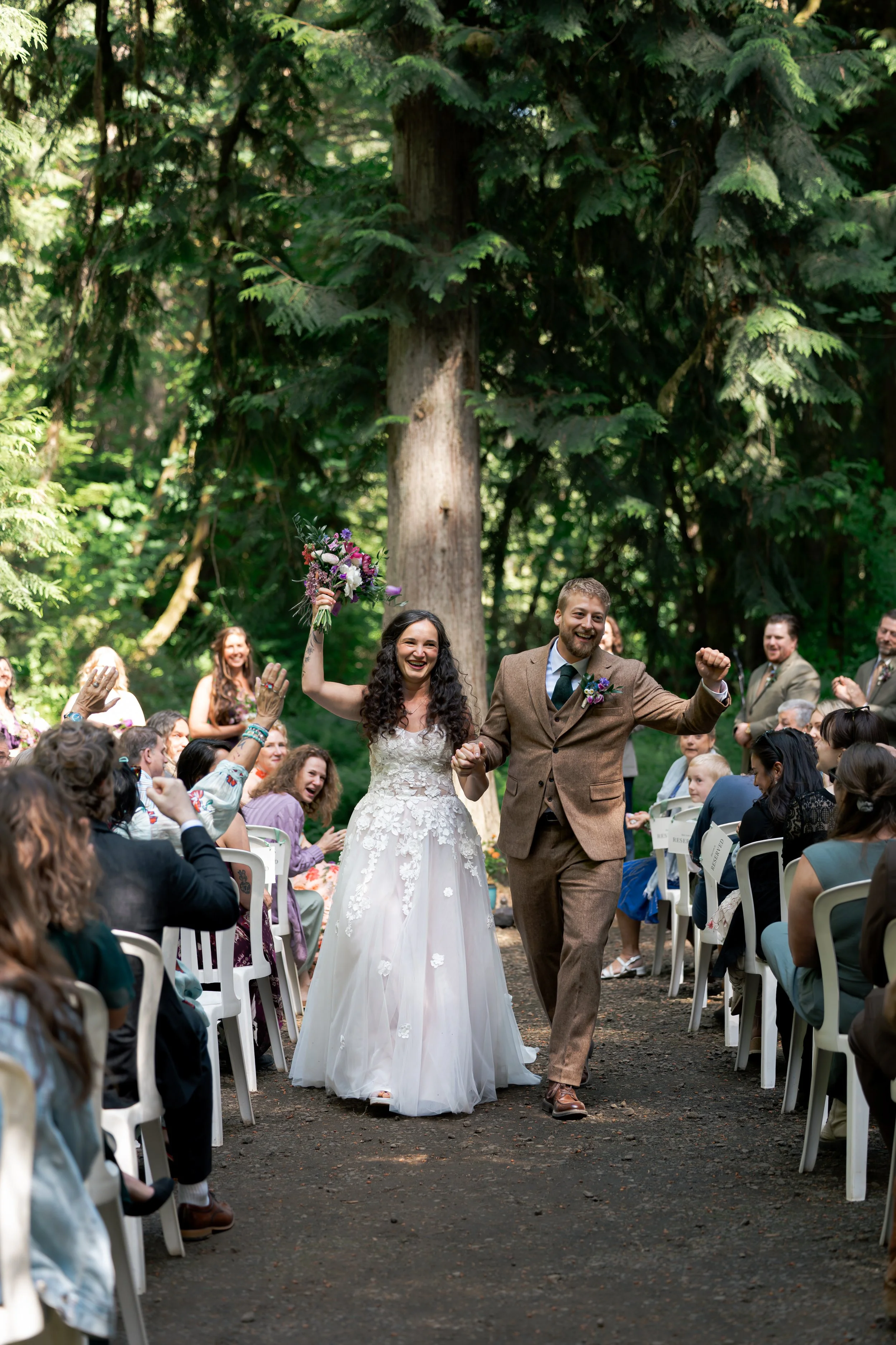 A bride and groom walk down the aisle at an outdoor wedding ceremony, surrounded by seated guests on either side. Forest Wedding Oregon Portland Wedding Photographer Pacific Northwest
