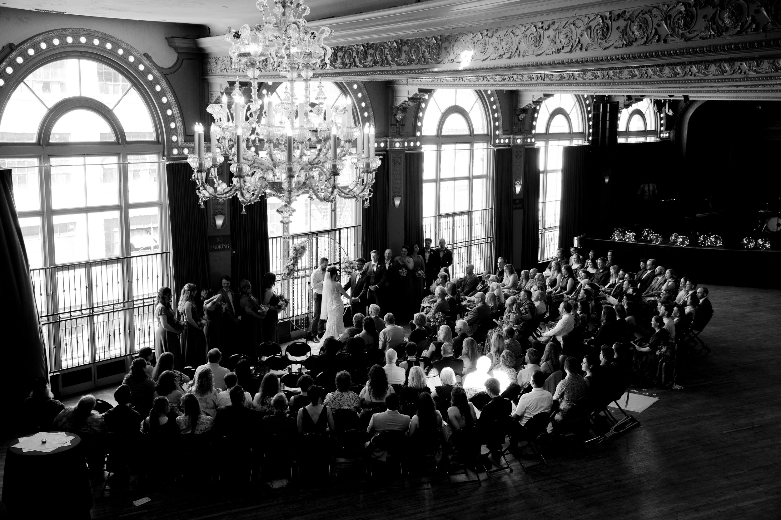 A black and white photo of a wedding ceremony in an elegant, ornate hall with large arched windows, a grand chandelier, and a gathered audience seated facing the couple. Luxury Oregon Wedding Photographer. Wedding Inspo. Elevated Moody Editorial