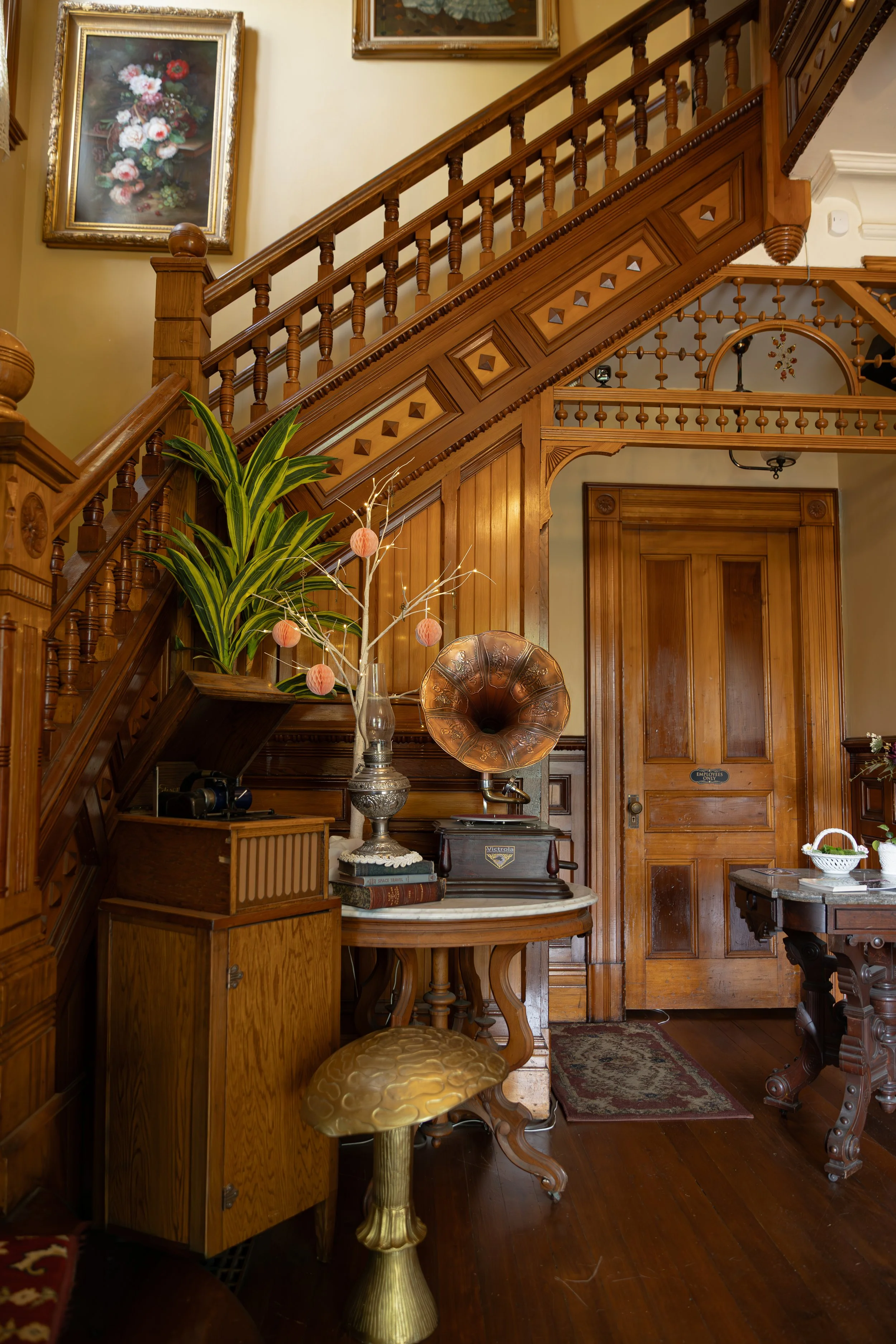 Interior of a home with wooden staircase, framed paintings, a large leafy plant, a vintage gramophone, a table with decorative items, and a door.