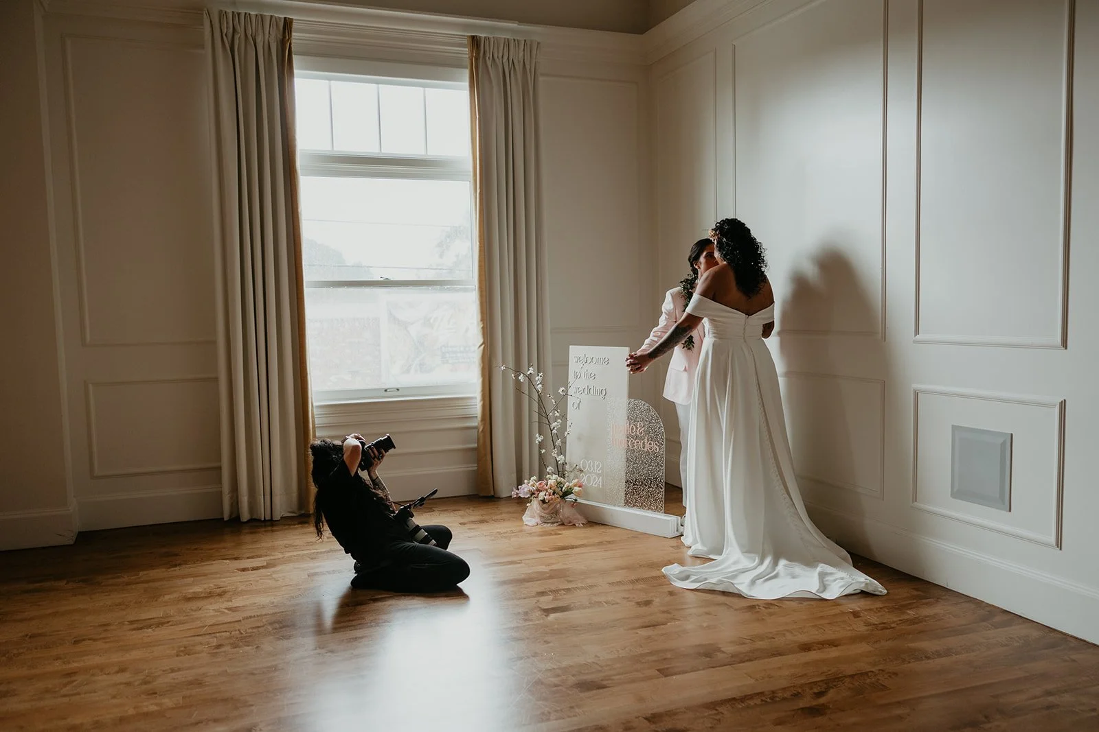 A bride in a white gown and a woman in pink are sharing a moment in a warmly lit room with wooden floors and white walls. Lauren Maye photographing a lesbian couple at the Arc Light Venue. A wedding sign and floral arrangements are also in the scene.