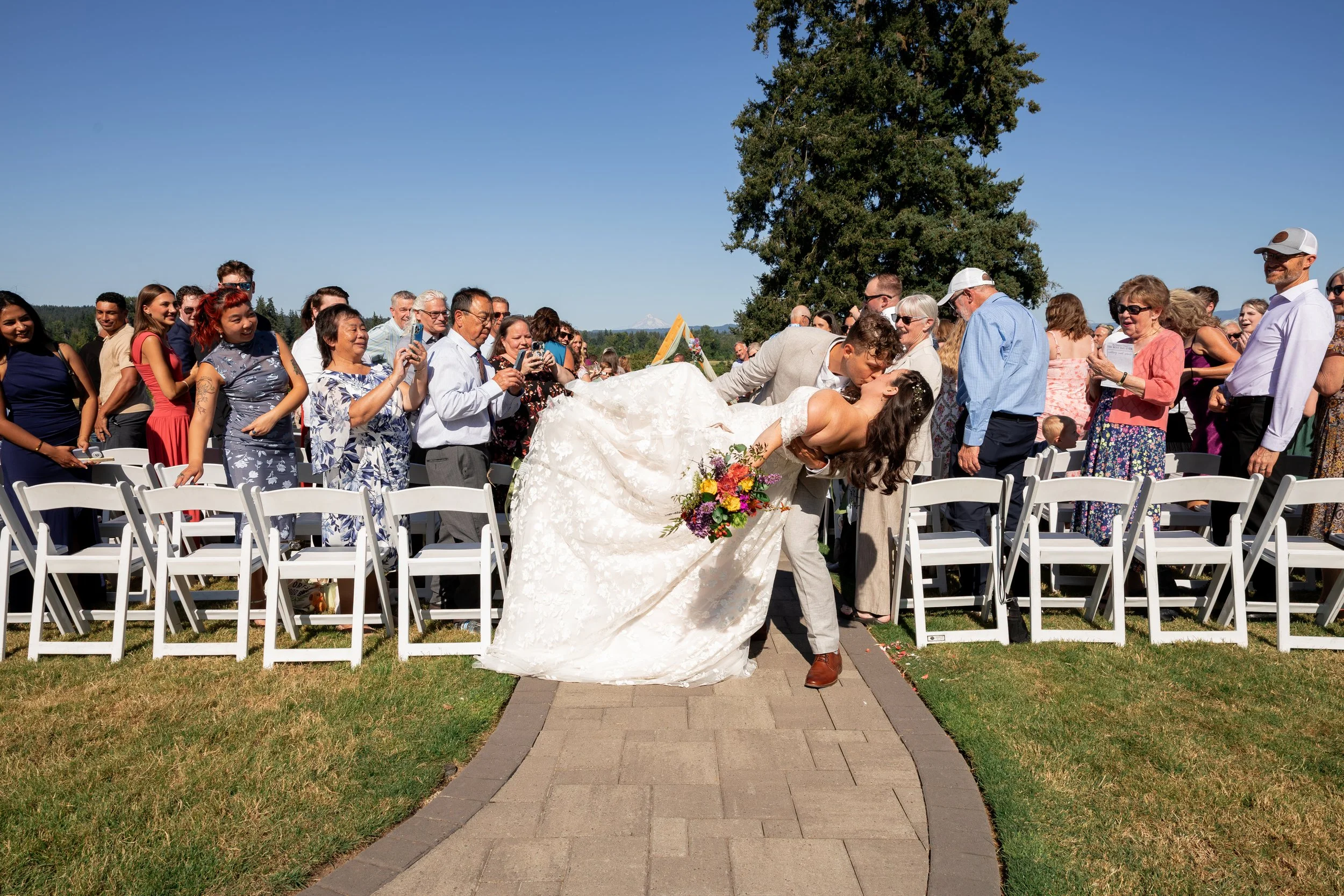 A bride and groom kissing at an outdoor wedding ceremony, surrounded by guests sitting on white chairs, with a large tree and clear blue sky in the background. Oregon wedding, summer wedding, best wedding photographer, Bay Area wedding, California