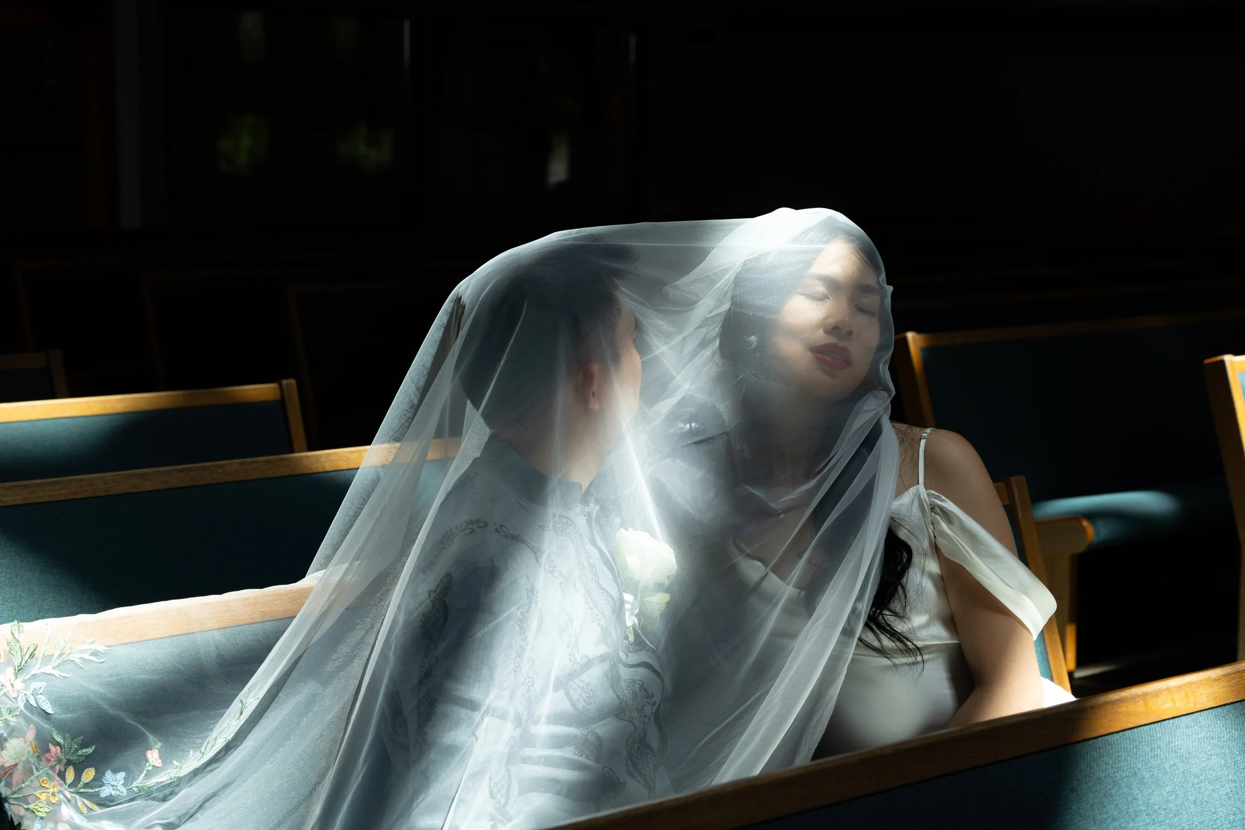 A bride wearing a white dress and veil sitting on a church pew with a woman leaning in to her, in a dark church with minimal lighting. Luxury Oregon Wedding Photographer. Wedding Inspo. Elevated Moody Editorial Church Wedding Natural light Sun beams