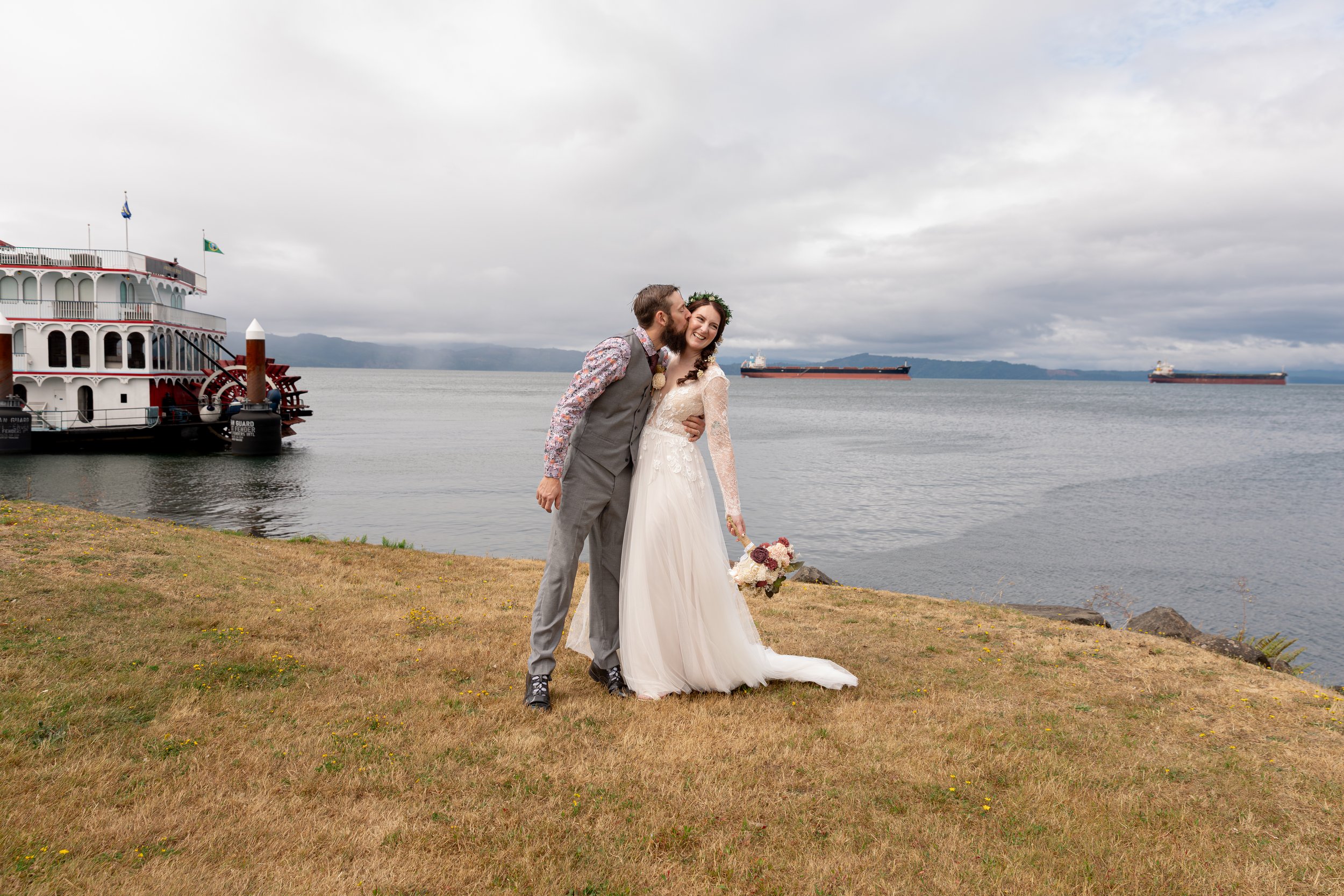 A newlywed couple sharing a tender moment by a body of water with a large boat docked nearby and a cargo ship in the distance, overcast sky. Oregon Coast, Oregon wedding, Bay Area wedding, California wedding, bridal portraits, whimsical wedding 