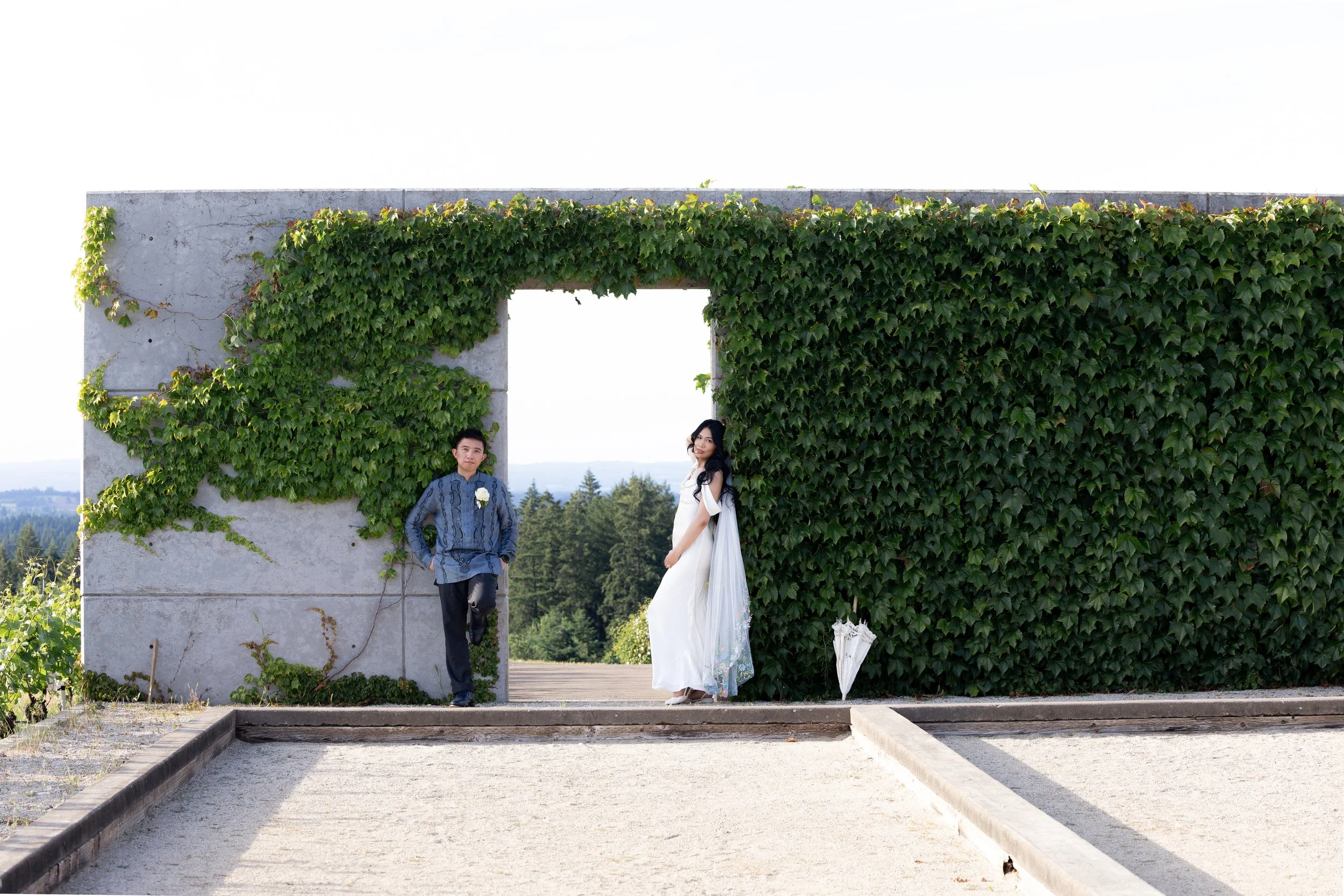 A man and woman standing in front of a large concrete wall covered with green ivy leaves, with a clear sky and trees in the background. Luxury Wedding Photographer. Vineyard Wedding. Oregon Wedding