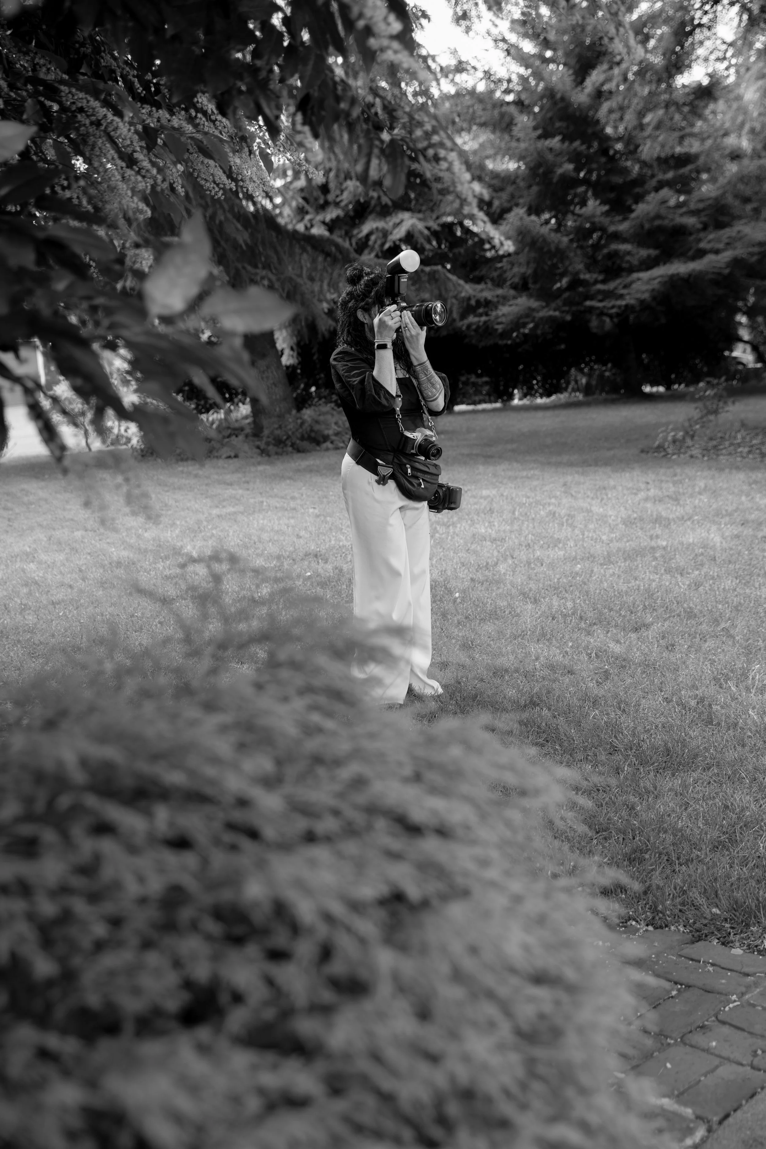 Lauren Maye at an Oregon City wedding, holding a camera and photographing the scene, with a film camera hanging from her neck, on a grassy area with bushes and trees in the background.