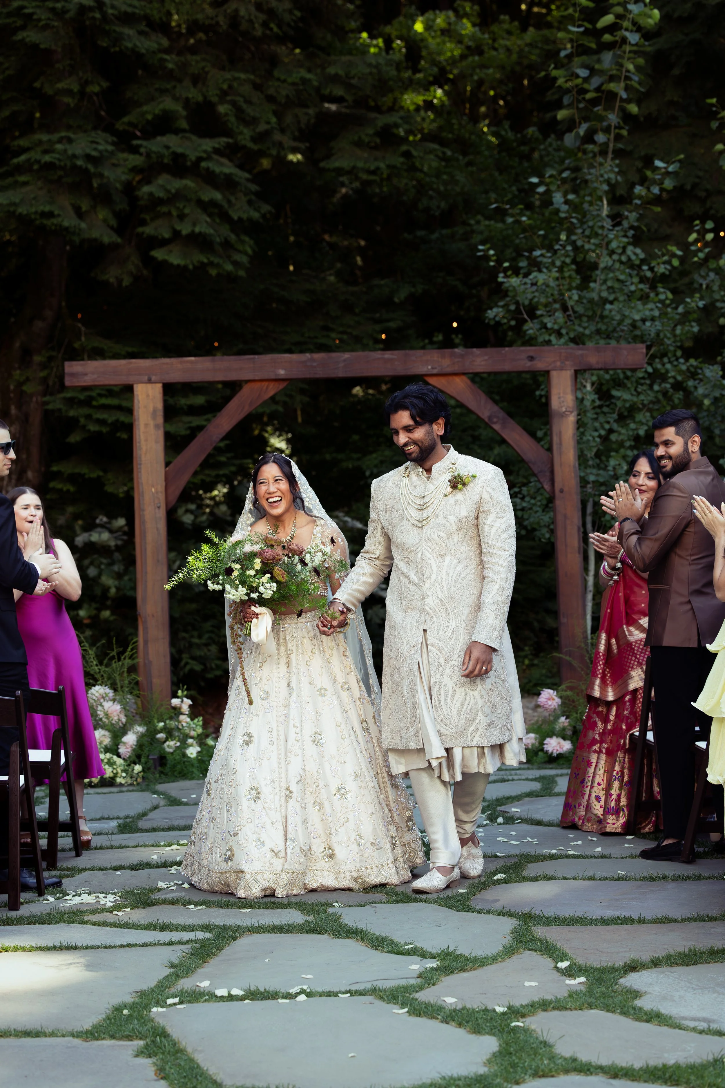 A wedding ceremony outdoors with a couple smiling and holding hands, surrounded by friends and family, set in a lush green garden with trees and floral decorations. Traditional Indian Wedding, Oregon wedding, California Wedding, Bay Area wedding,