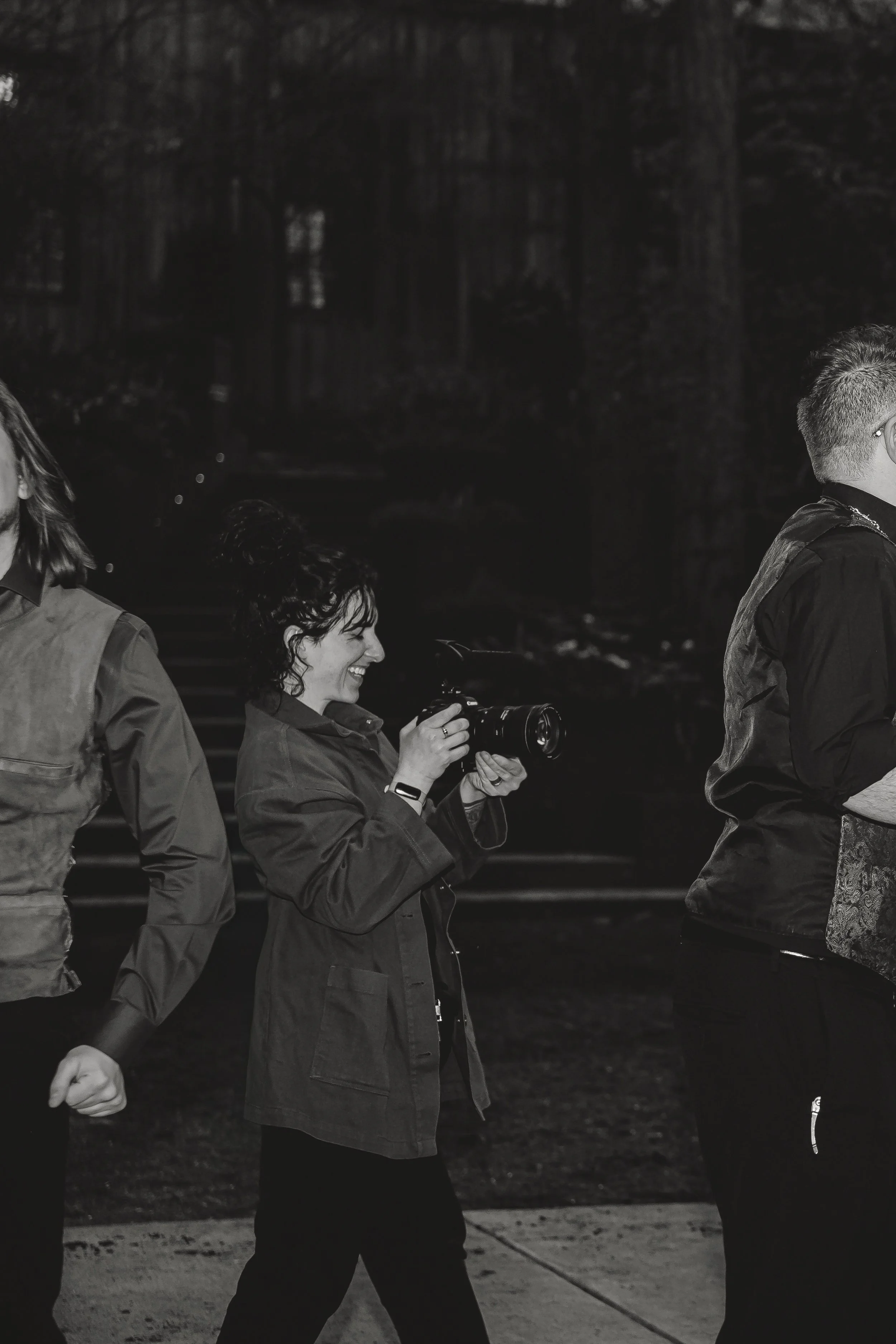 People walking outdoors at night, one person smiling and taking photos, others partially visible. Lauren Maye taking video at a queer wedding at Camp Colton in Oregon.
