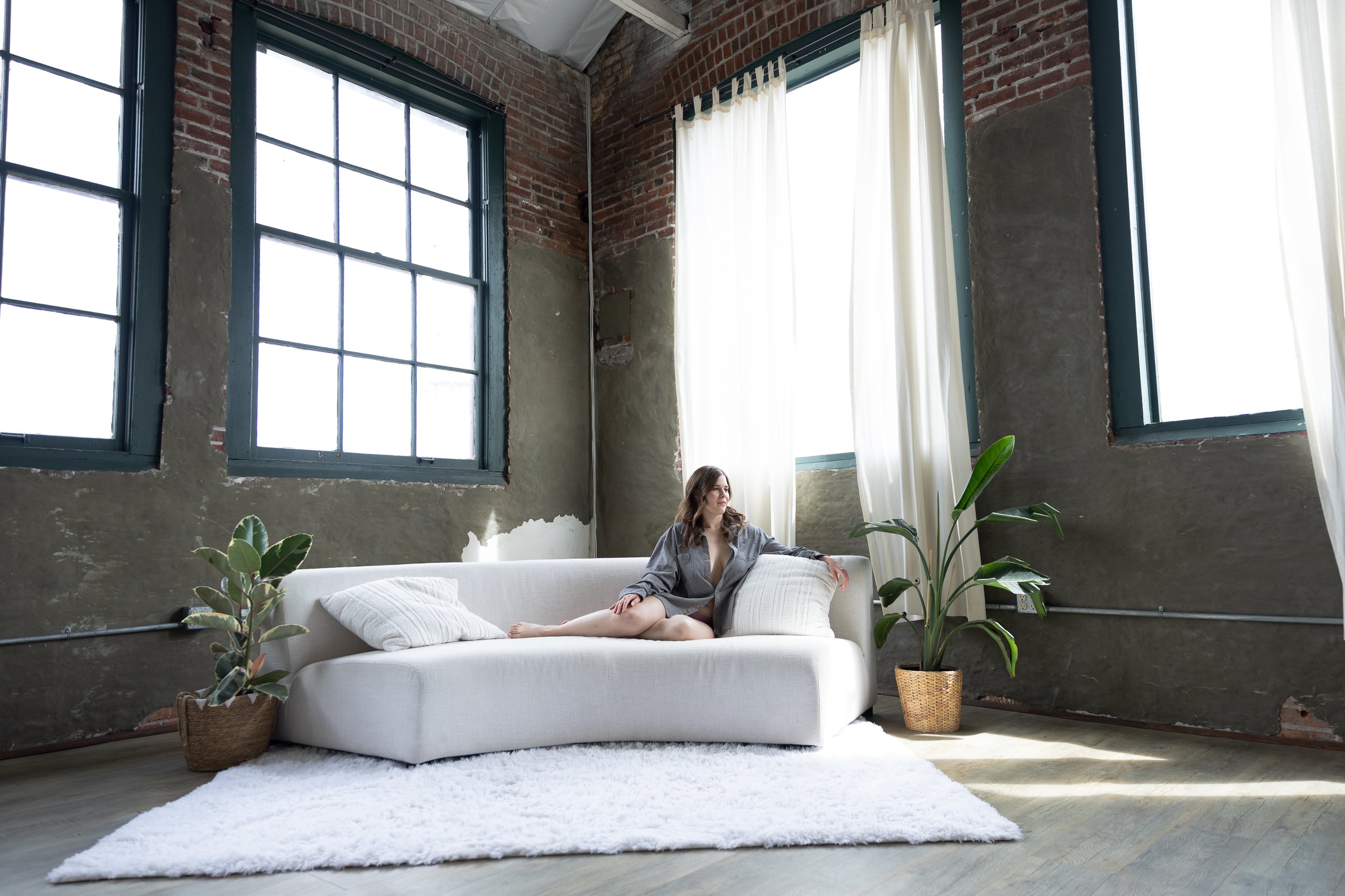 A woman sitting on a white sofa in a loft-style room with large windows, peeling paint walls, and green houseplants.