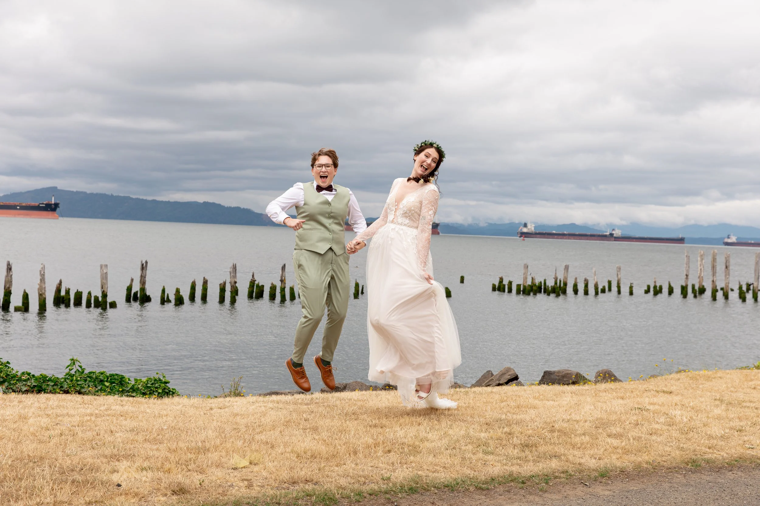 A couple dressed in wedding attire, with the groom jumping in the air, standing near a body of water with ships in the background under an overcast sky. Oregon Coast Wedding, Best wedding photographer in Oregon Best wedding photographer in California