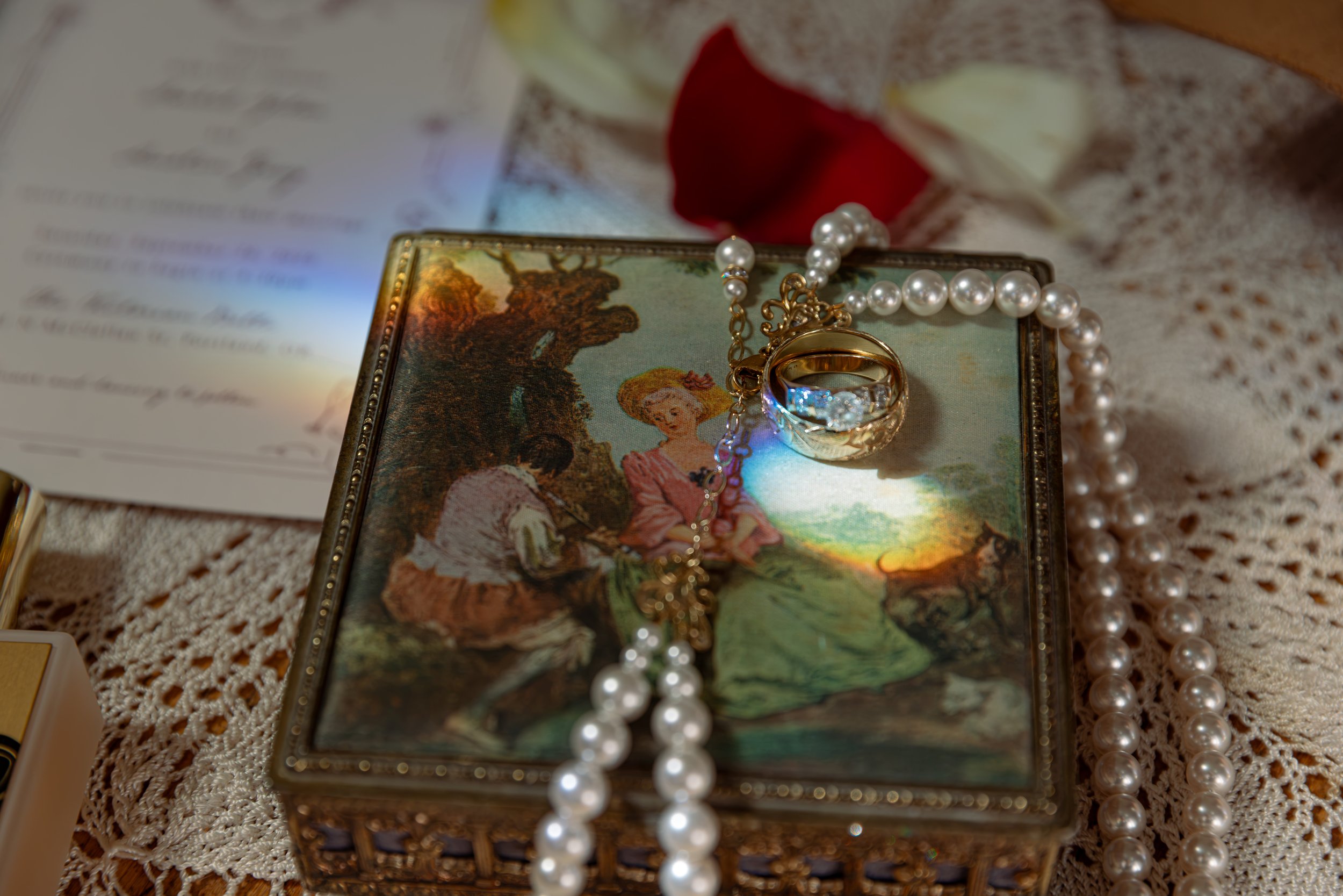 A string of pearl jewelry placed on a decorative picture frame featuring a painting of a woman and a man sitting outdoors under a tree, with a red flower and a candle on a lace tablecloth.