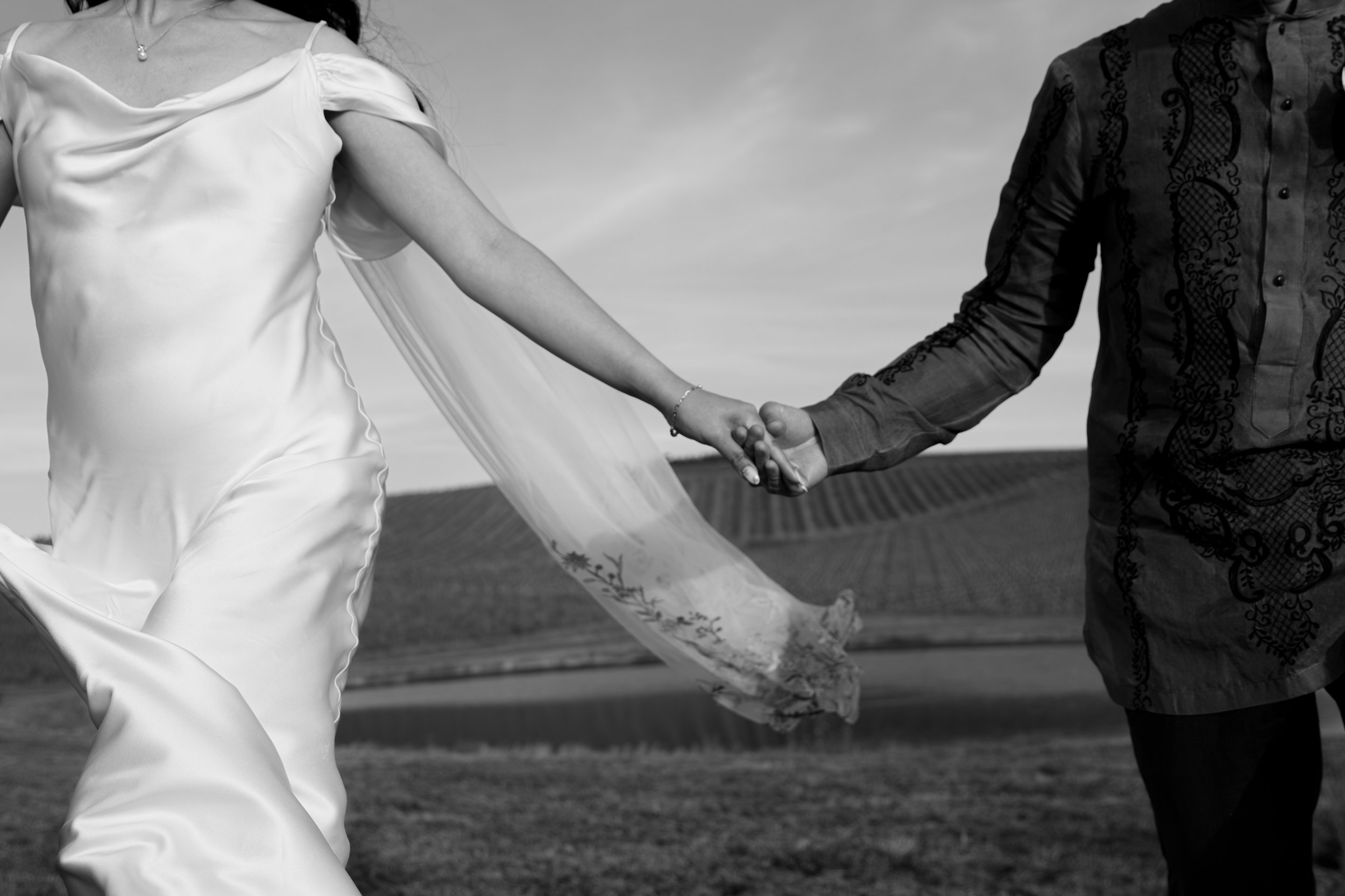 A couple holding hands outdoors, with the woman wearing a wedding dress and the man wearing a patterned shirt, against a cloudy sky background. Luxury Oregon Wedding Photographer. Wedding Inspo. Elevated Moody Editorial