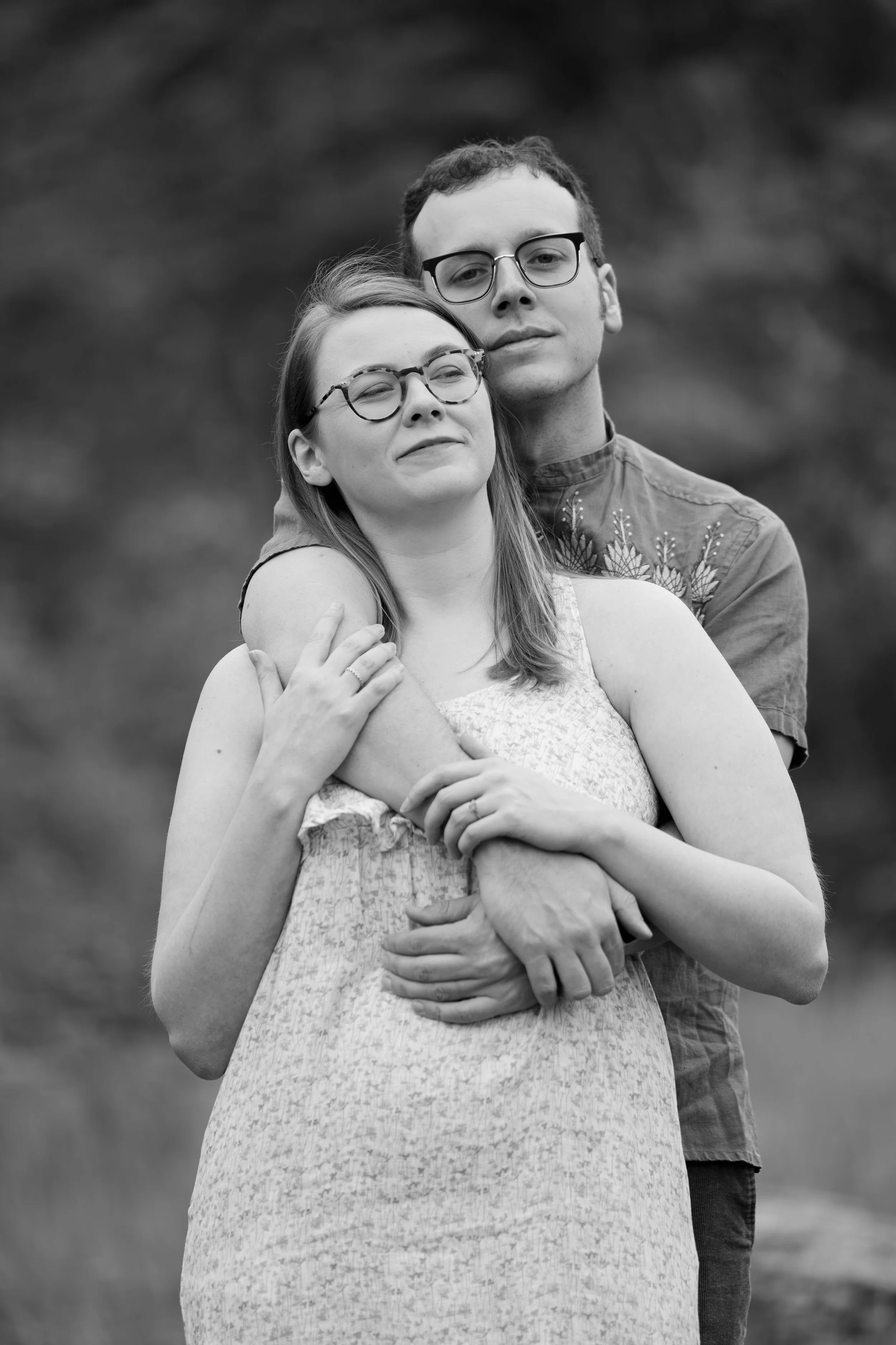 A black-and-white photo of a couple standing outdoors. The woman has long hair, glasses, and is wearing a sleeveless dress. The man, wearing glasses and a shirt with embroidery, stands behind her with his arms around her. Both are showing relaxed exp