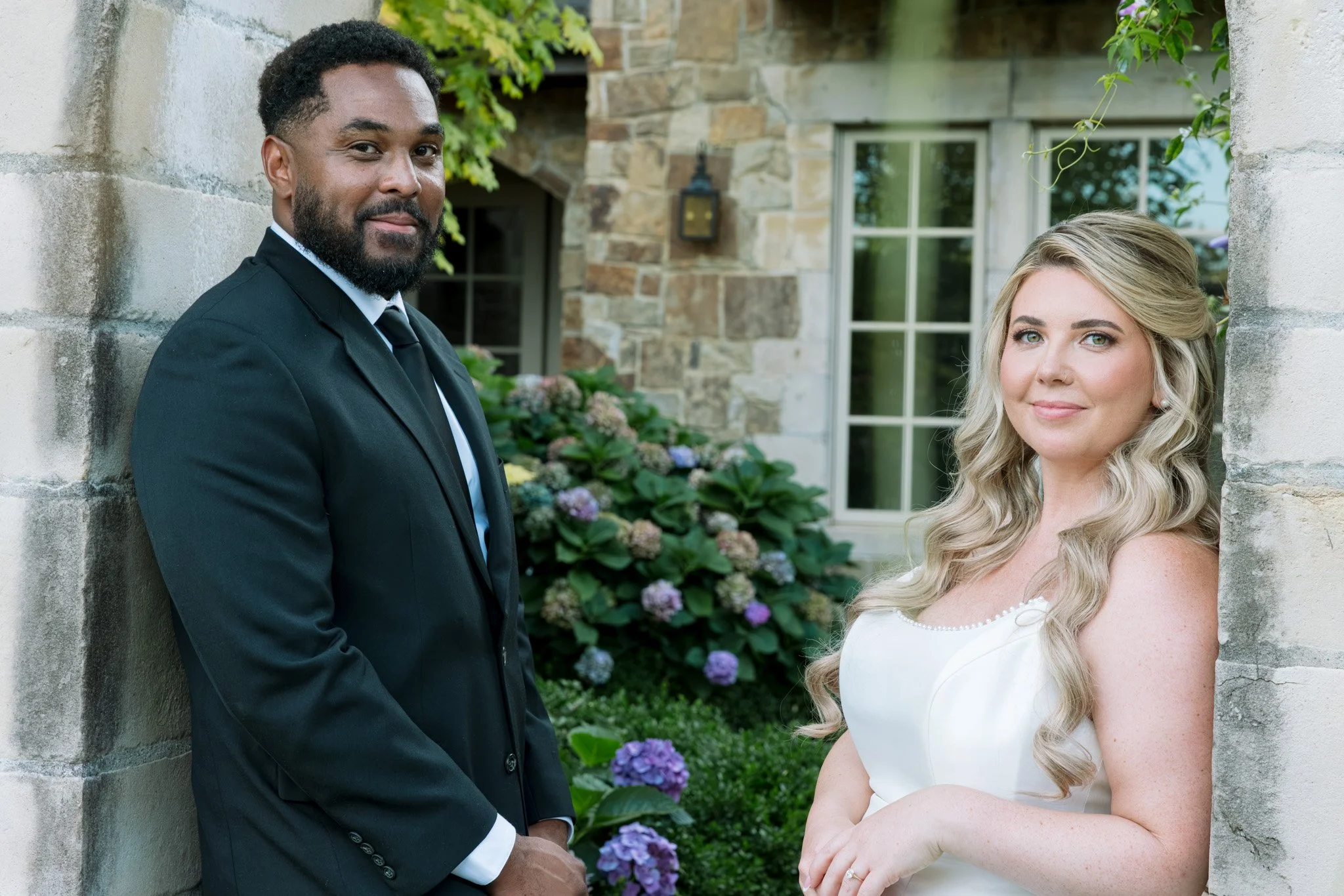 A man in a black suit and a woman in a white dress posing outdoors near a stone wall and a window, with greenery and purple flowers in the background. Chateau wedding Bay Area Wedding California Wedding Oregon Wedding Pacific Northwest Wedding White