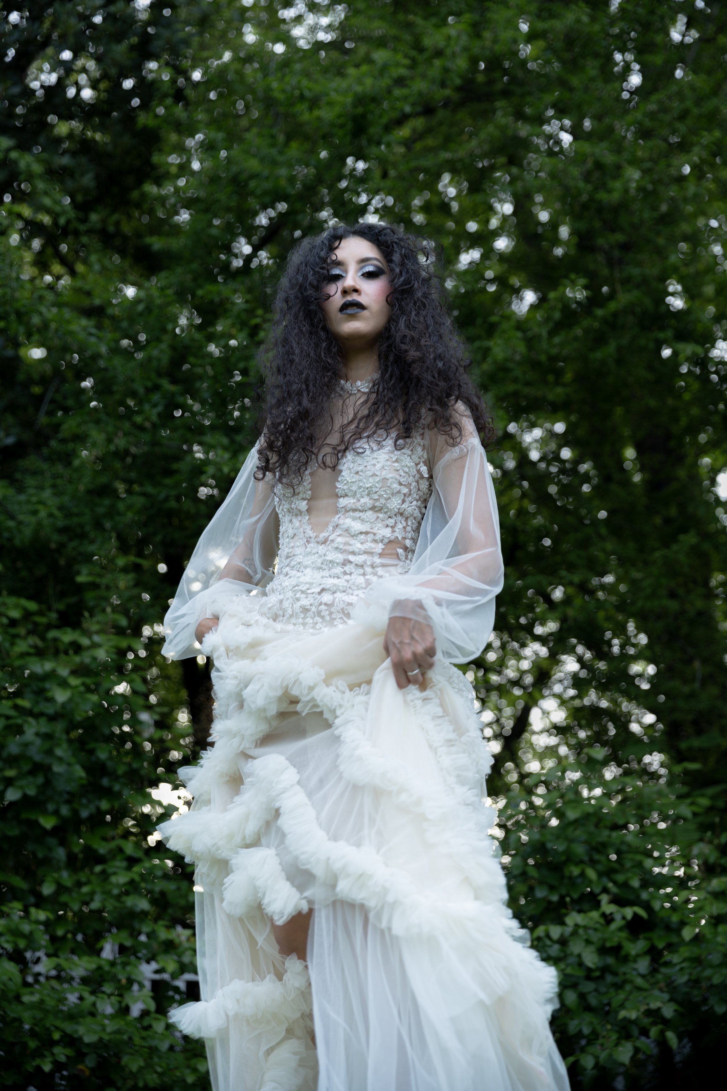 A person with dark, curly hair and dark makeup wearing a white, gothic-style lace and tulle dress standing outdoors among green foliage. Luxury Oregon Wedding Photographer. Wedding Inspo. Elevated Moody Editorial