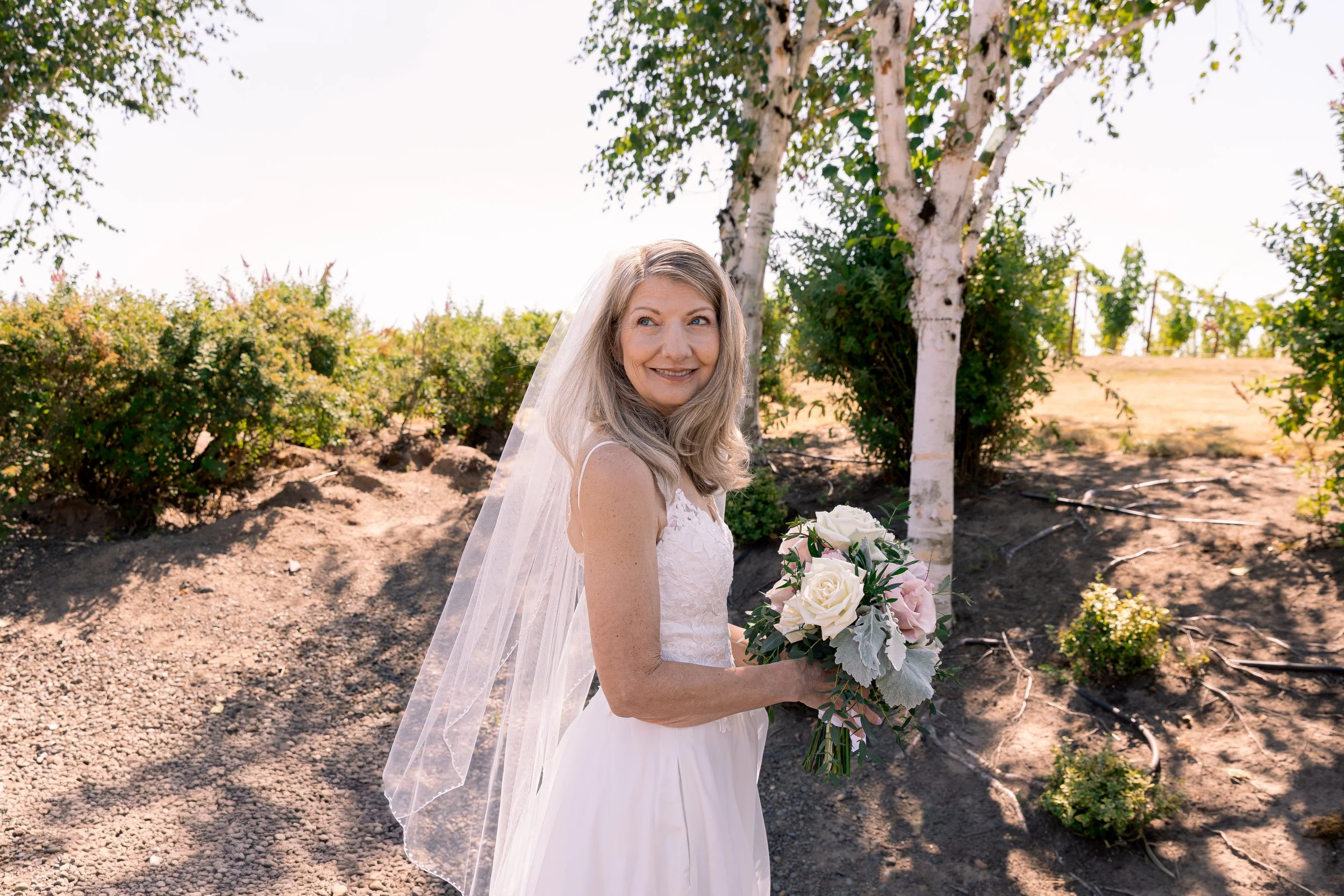 A smiling woman in a white wedding dress holding a bouquet of white and pink roses, standing outdoors in front of trees and bushes. Bridal portraits, oregon wedding california wedding bay area wedding portland wedding pacific northwest wedding