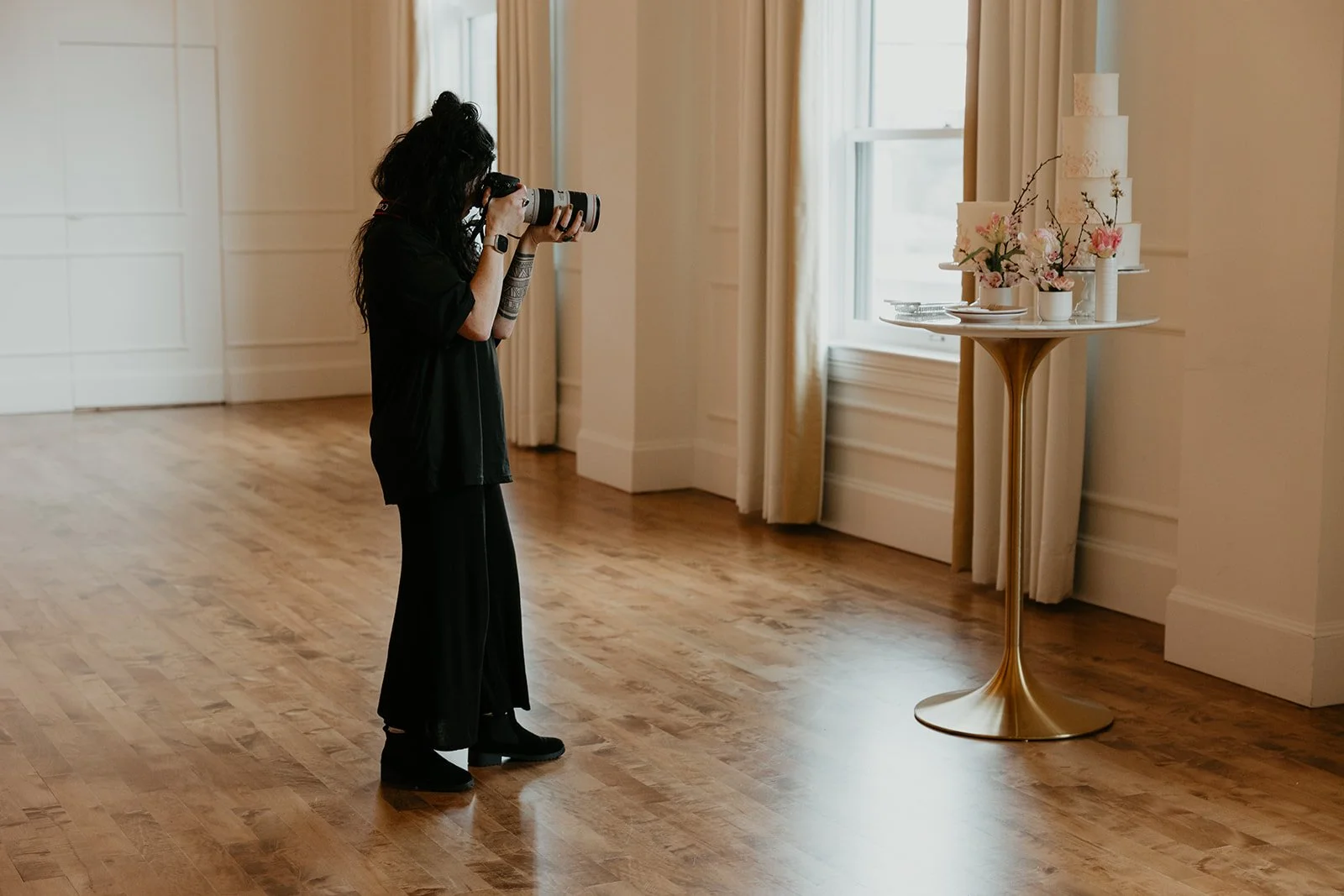 A woman taking a photograph of a wedding cake on a round gold stand in a well-lit room with wooden floors, white walls, and large windows with beige curtains. Lauren Maye at the Arc Light Venue in Oregon City at a styled shoot