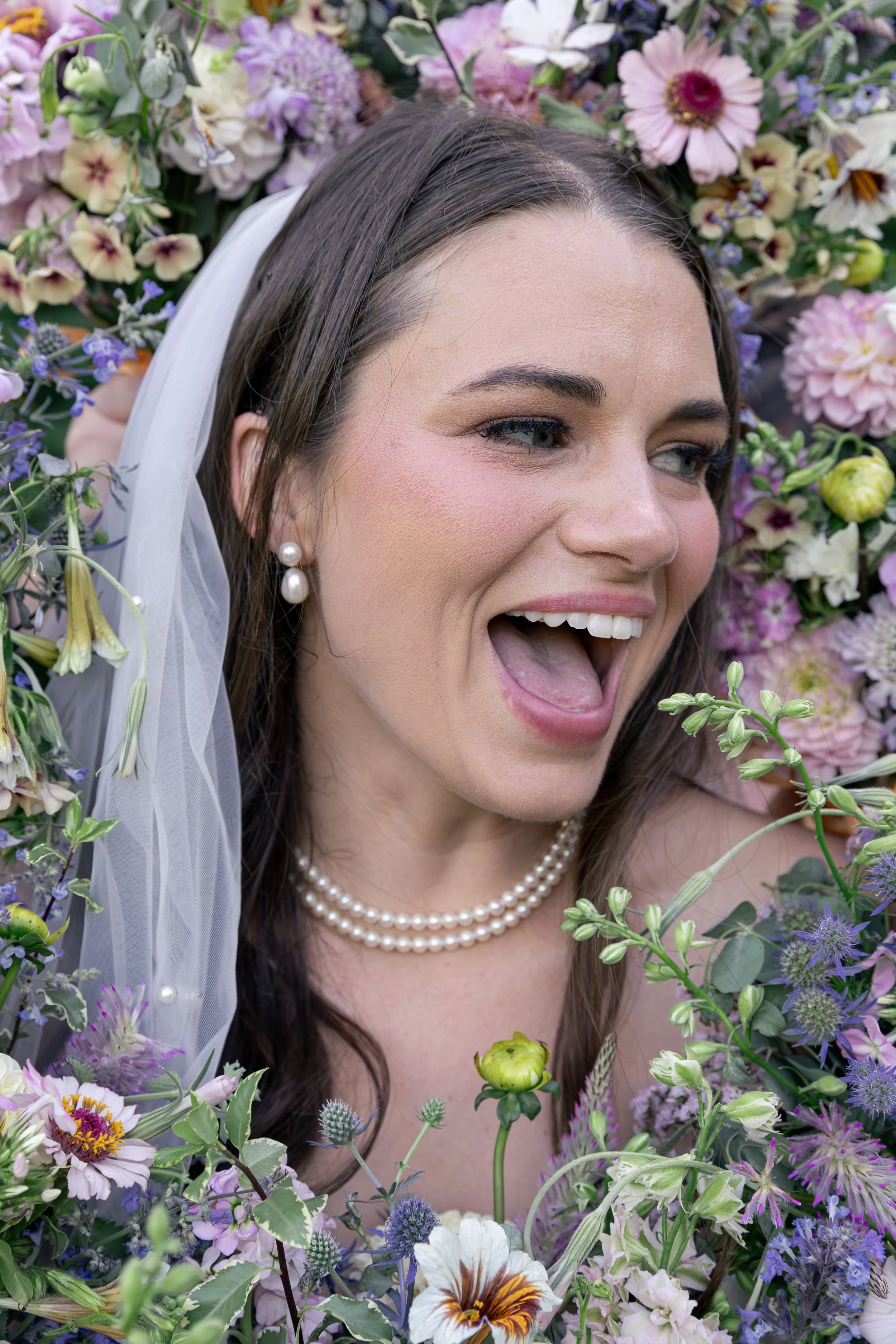 Close-up of a woman with long dark hair, wearing pearl earrings and necklace, smiling with her tongue out, surrounded by colorful flowers and greenery. Bridal portrait inspo. Oregon + Travel Wedding photographer. Best wedding photographer in Oregon. 