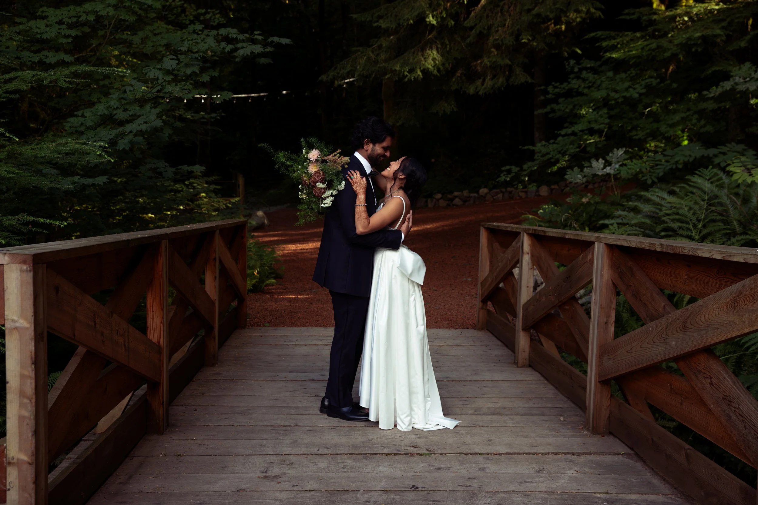 Bride and groom standing together on a wooden bridge in a forest, embracing and smiling at each other, with the bride holding a bouquet of flowers. Oregon Wedding, California Wedding, Bay Area Wedding, Pacific Northwest Wedding, Portland Wedding