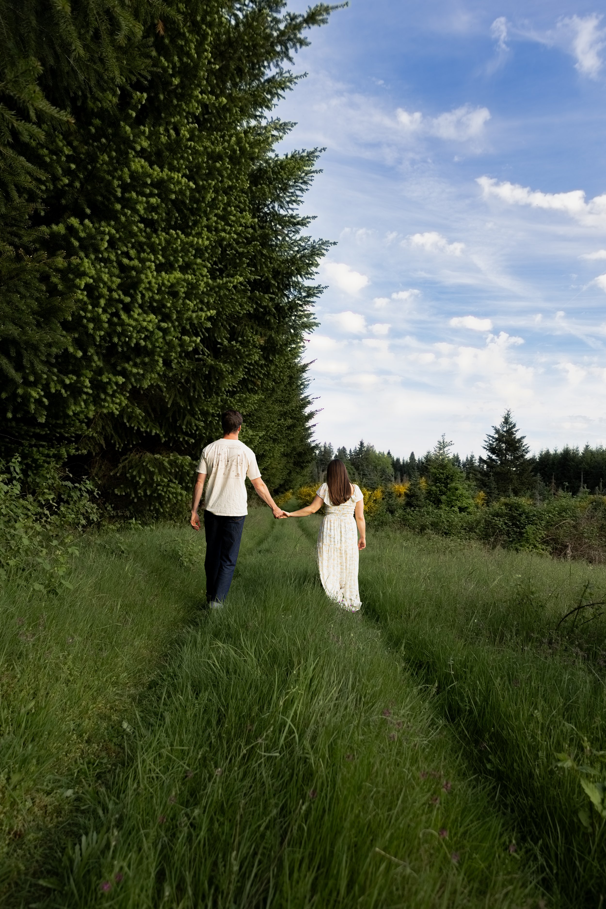 A couple holding hands walking along a grassy path through a forested area with tall trees and a partly cloudy sky.