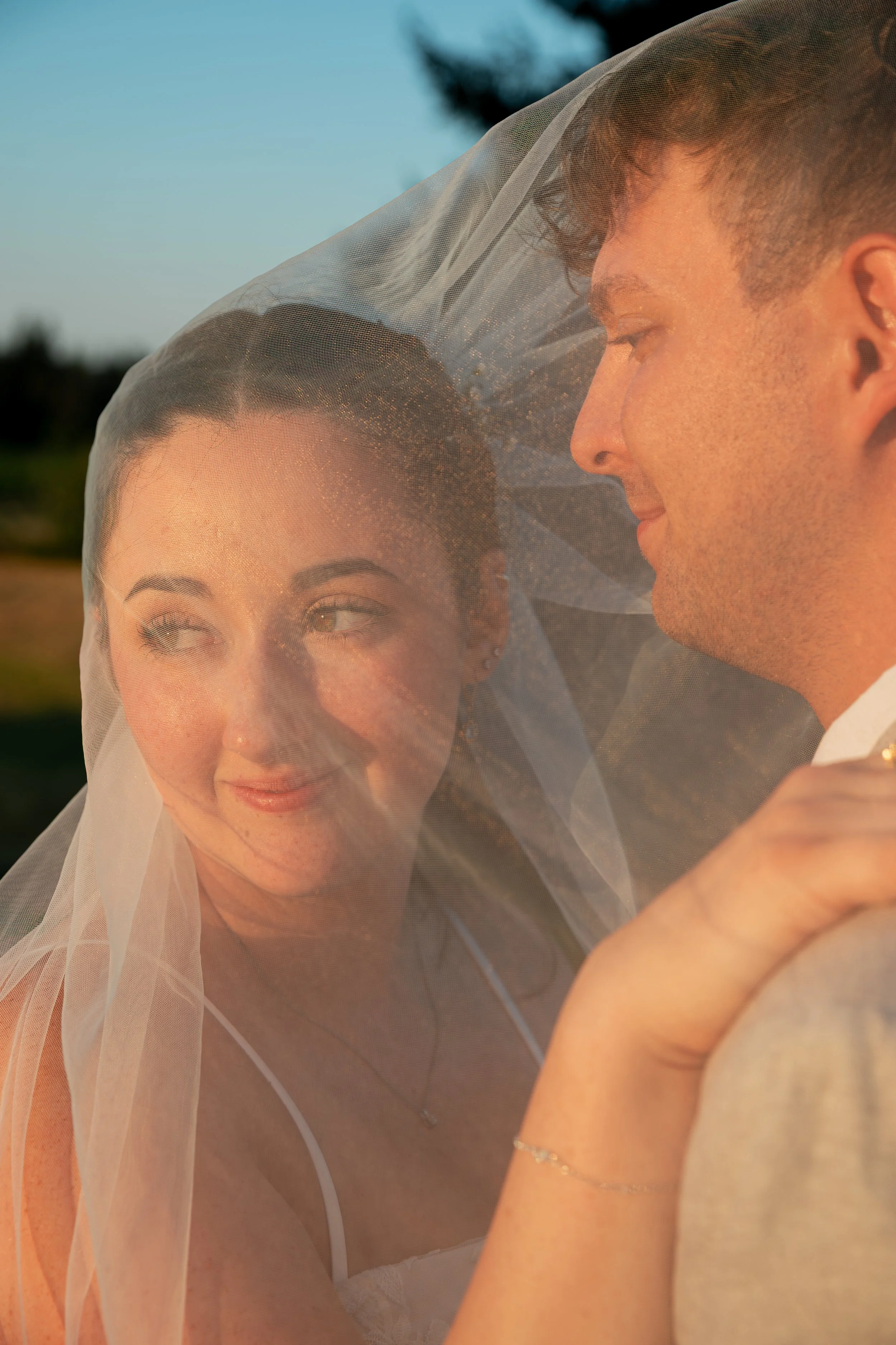 A close-up of a couple embracing outdoors, with the woman wearing a sheer white veil and the man smiling at her, both illuminated by warm sunlight. Chateau wedding Bay Area Wedding California Wedding Oregon Wedding Pacific Northwest Wedding 