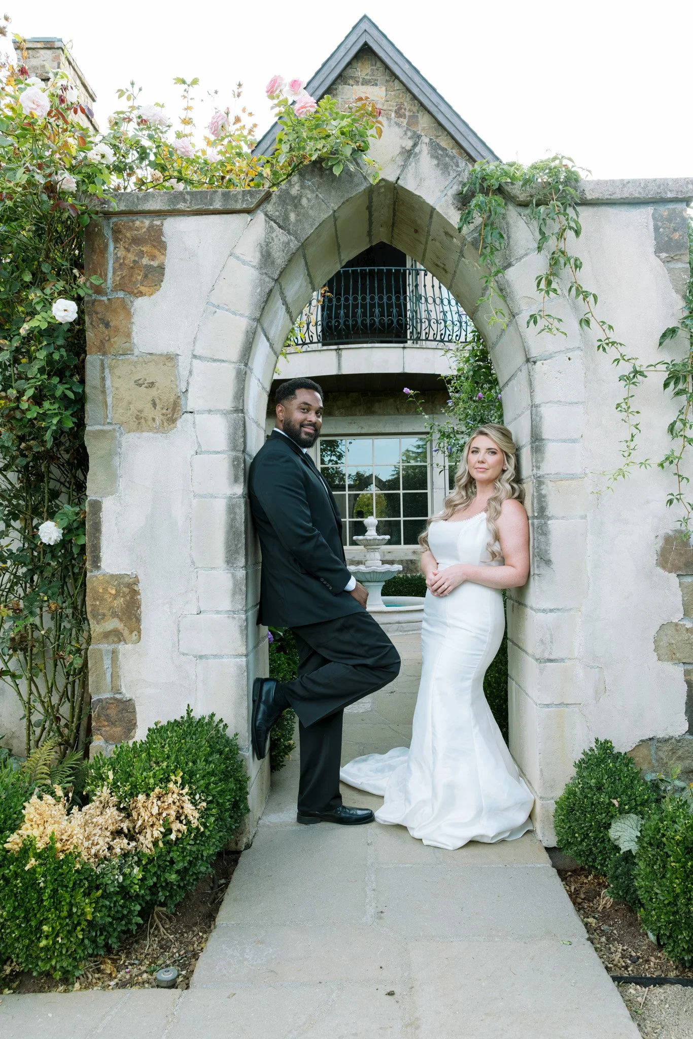 A bride and groom are standing under an arched stone entryway decorated with climbing plants and flowers, with a two-story house and garden in the background. Luxury wedding photography, Portland wedding, Oregon Wedding, Bay Area Wedding