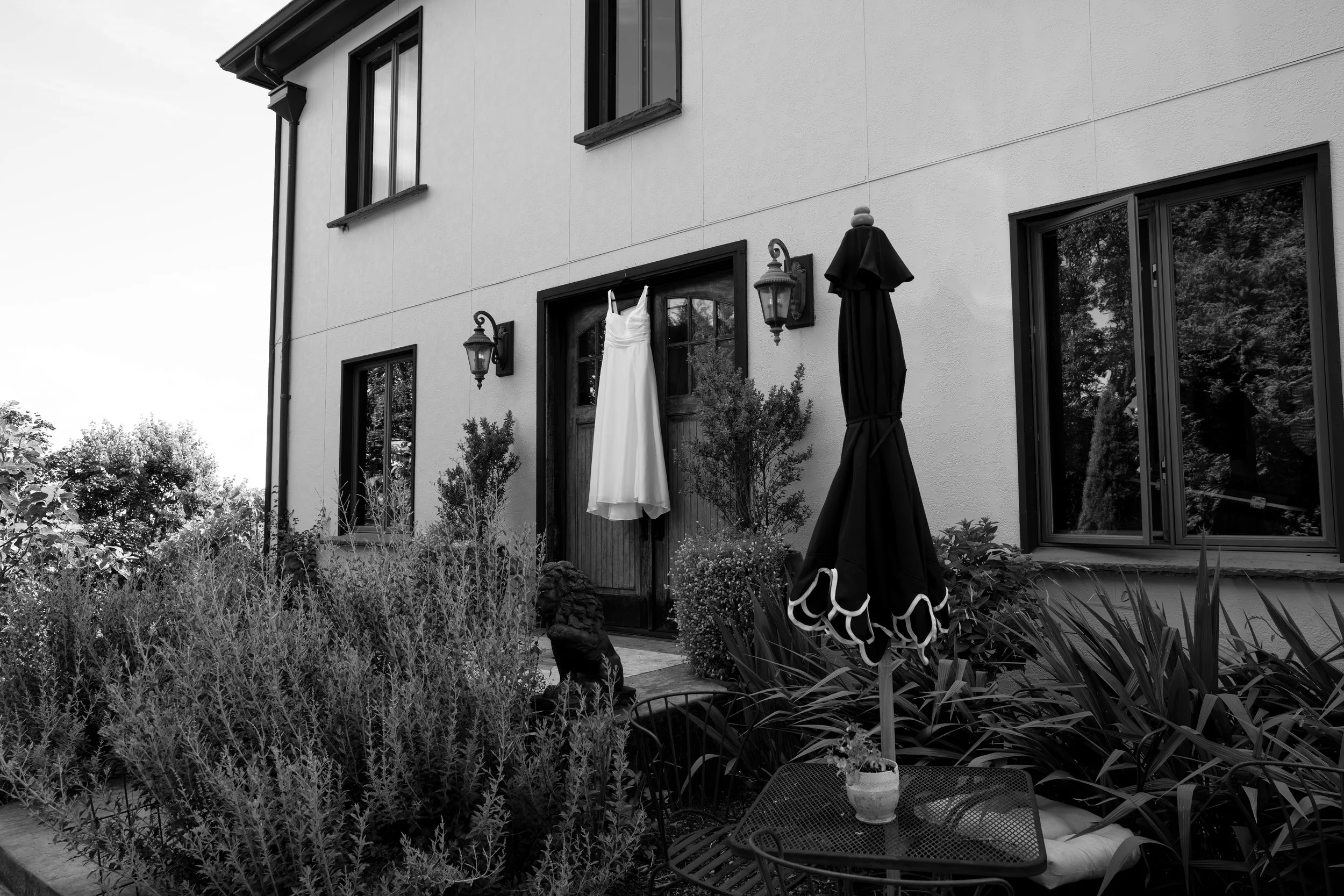 Black and white photo of a house exterior with a garden. There is a white dress hanging on a door, two outdoor lamps, and an umbrella over a table with chairs.