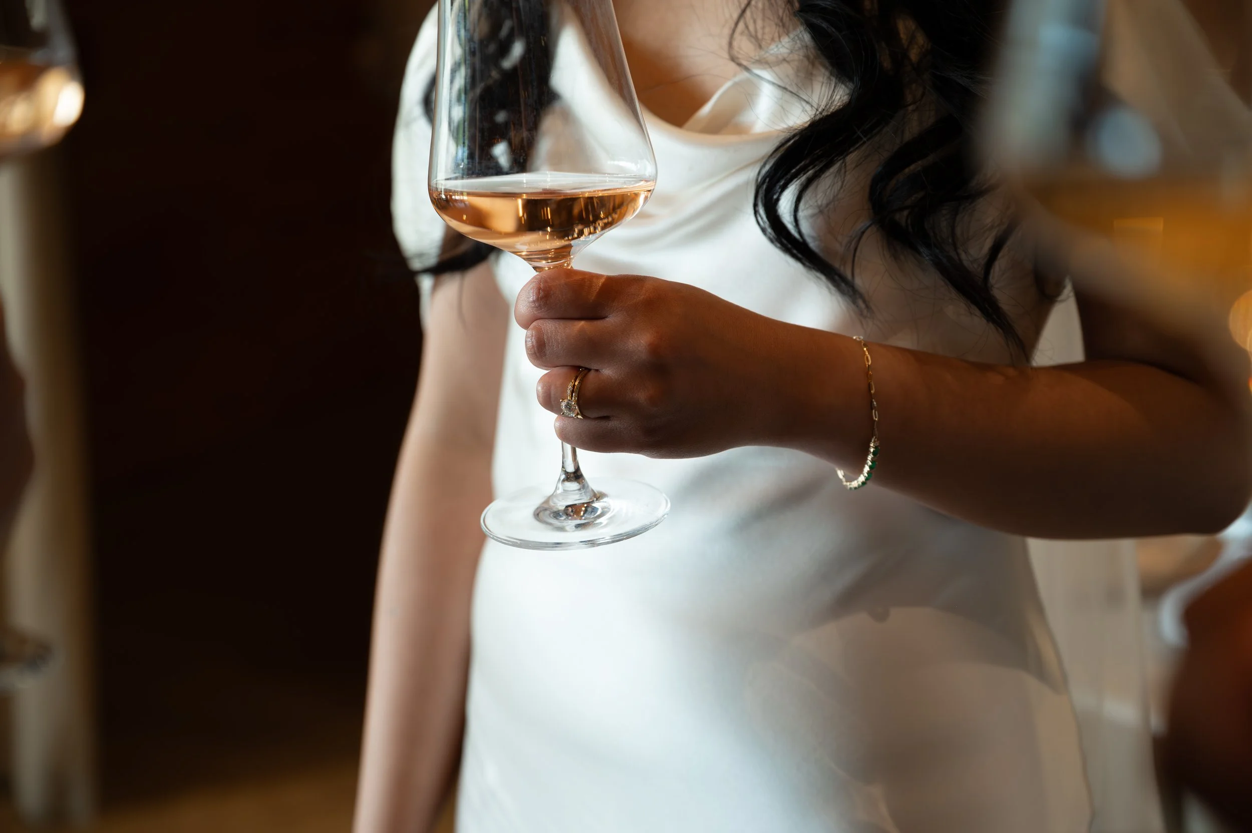 A woman with dark hair wearing a white dress is holding a glass of rosé wine in her right hand. She is wearing a ring on her ring finger and a bracelet on her wrist. The background appears to be a warm, softly lit setting.