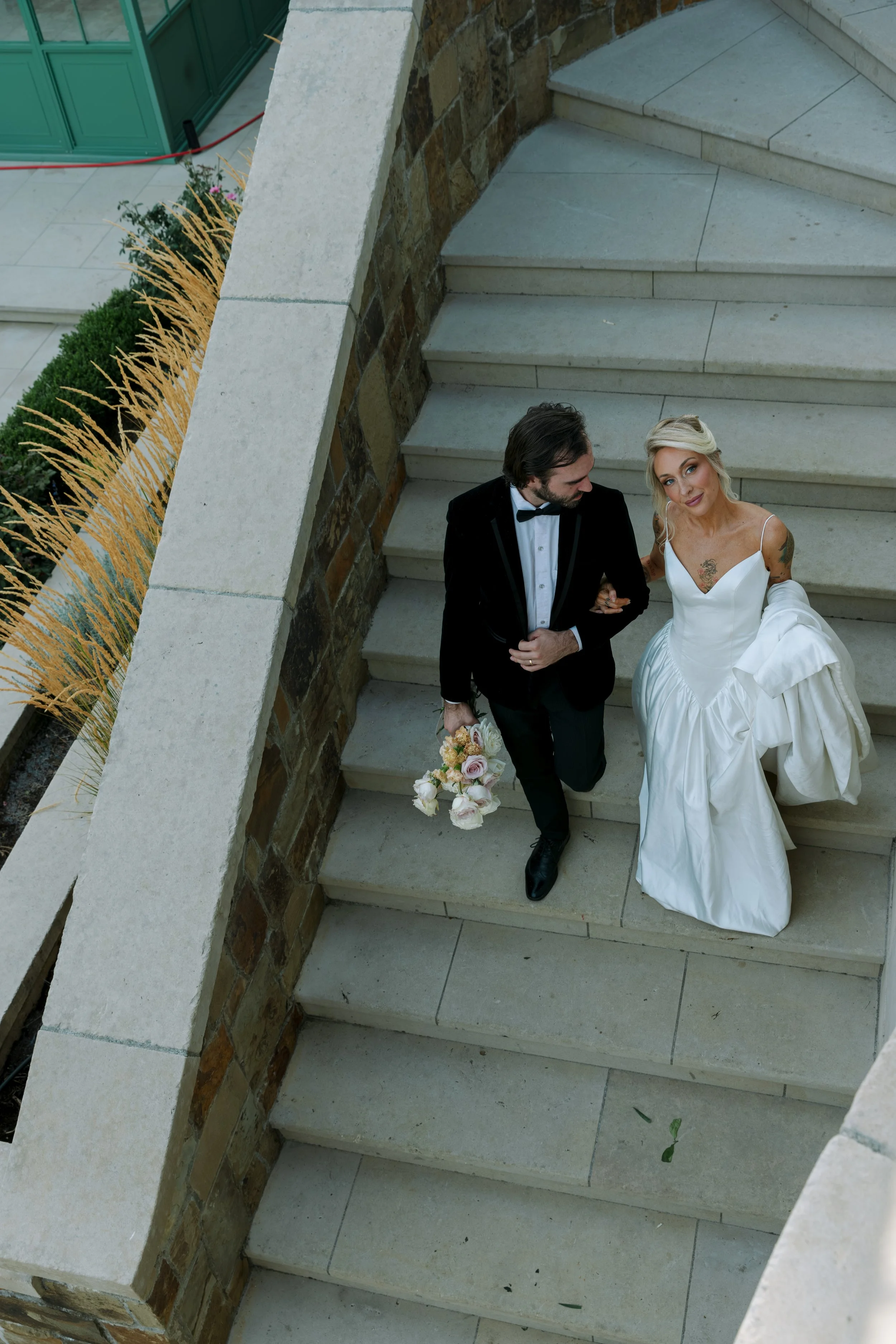 A bride and groom posing on outdoor stairs. The groom is wearing a black tuxedo with a bow tie, holding a bouquet of flowers. Chateau wedding Bay Area Wedding California Wedding Oregon Wedding Pacific Northwest Wedding White dress Bridal Portraits 