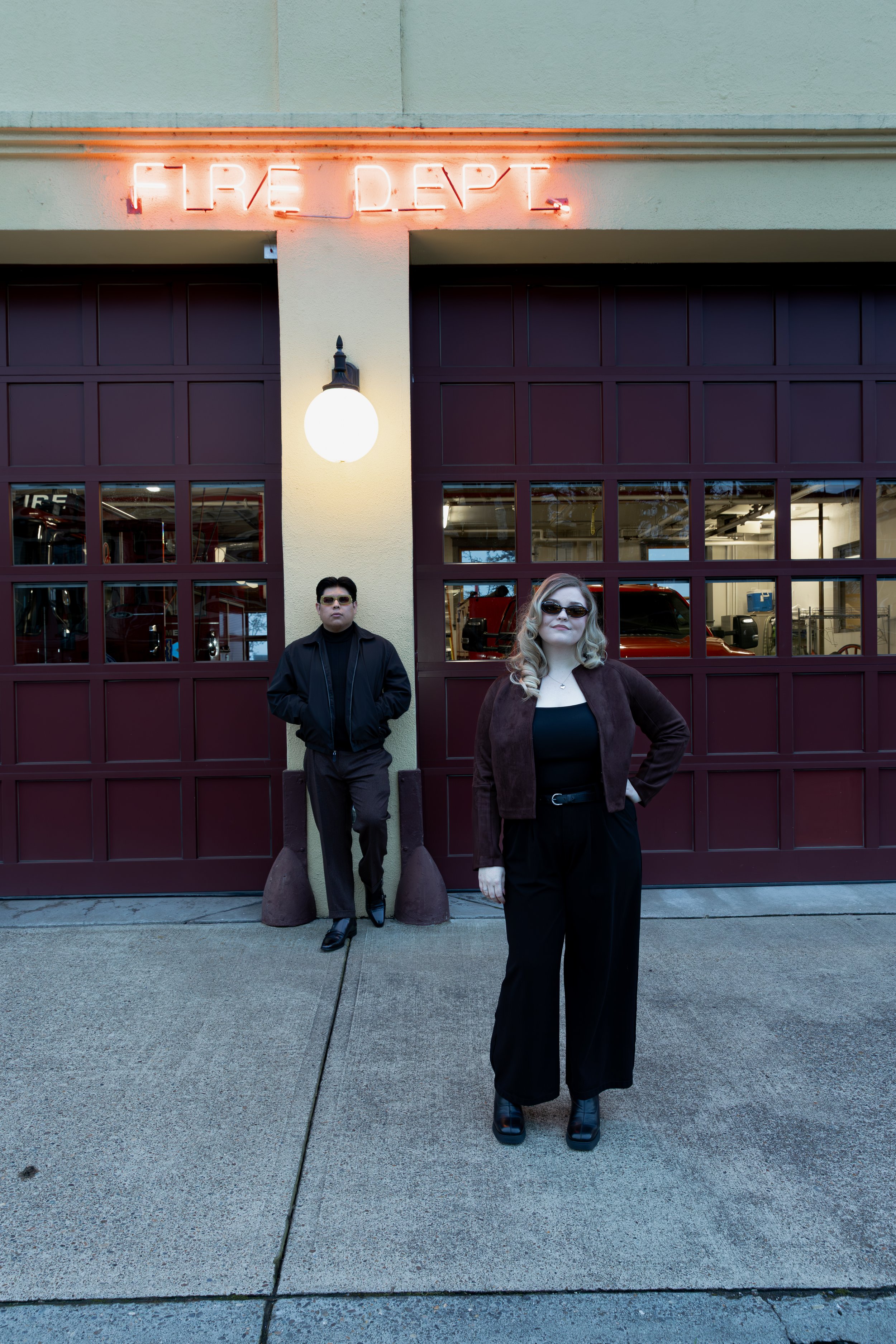 Two individuals stand outside a building with a maroon garage door and a glowing orange sign that reads "Fire Dept." in neon. Luxury Oregon Wedding photographer. Engagement Photo inspiration. city urban Engagement Photos
